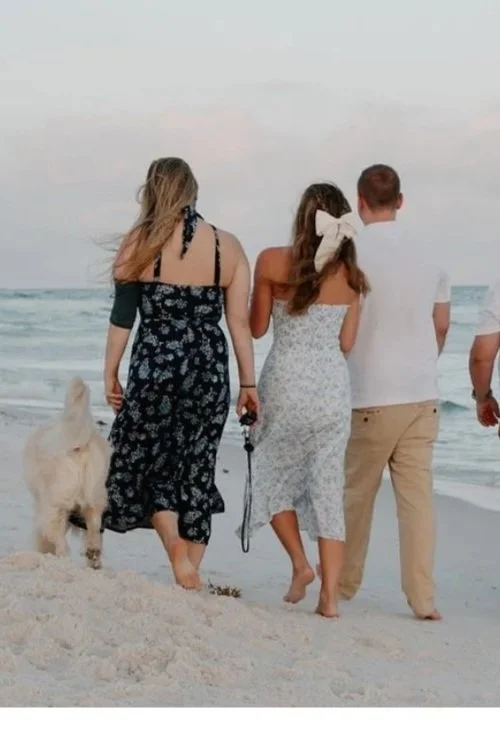 Four people and a dog walking on the beach, seen from the back, with the ocean and sky in the background.