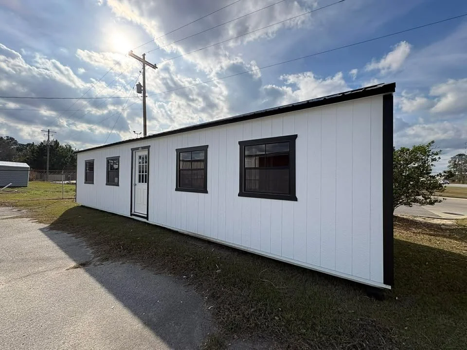A white rectangular mobile home with black trim around three windows and a door, situated outdoors on a grassy area with a partly cloudy sky overhead.