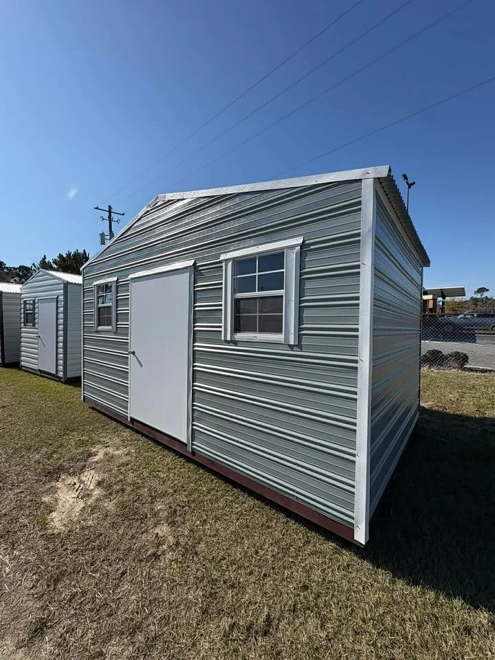 A metal shed with a white door and two windows, situated on grass with clear blue sky overhead.
