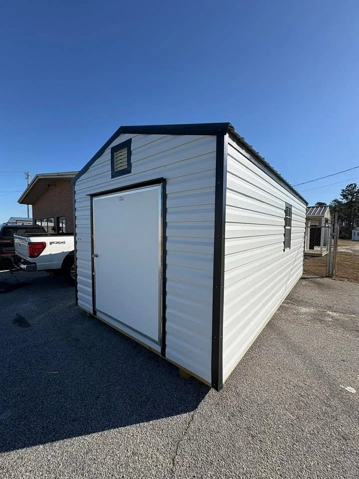 White metal storage shed with black trim, a large door, and a small vent at the gable end, situated on an asphalt driveway in a residential area.