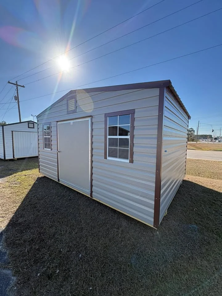 Metal storage shed with two small windows and a sliding door, situated on a grassy lot under a clear blue sky with bright sunlight.