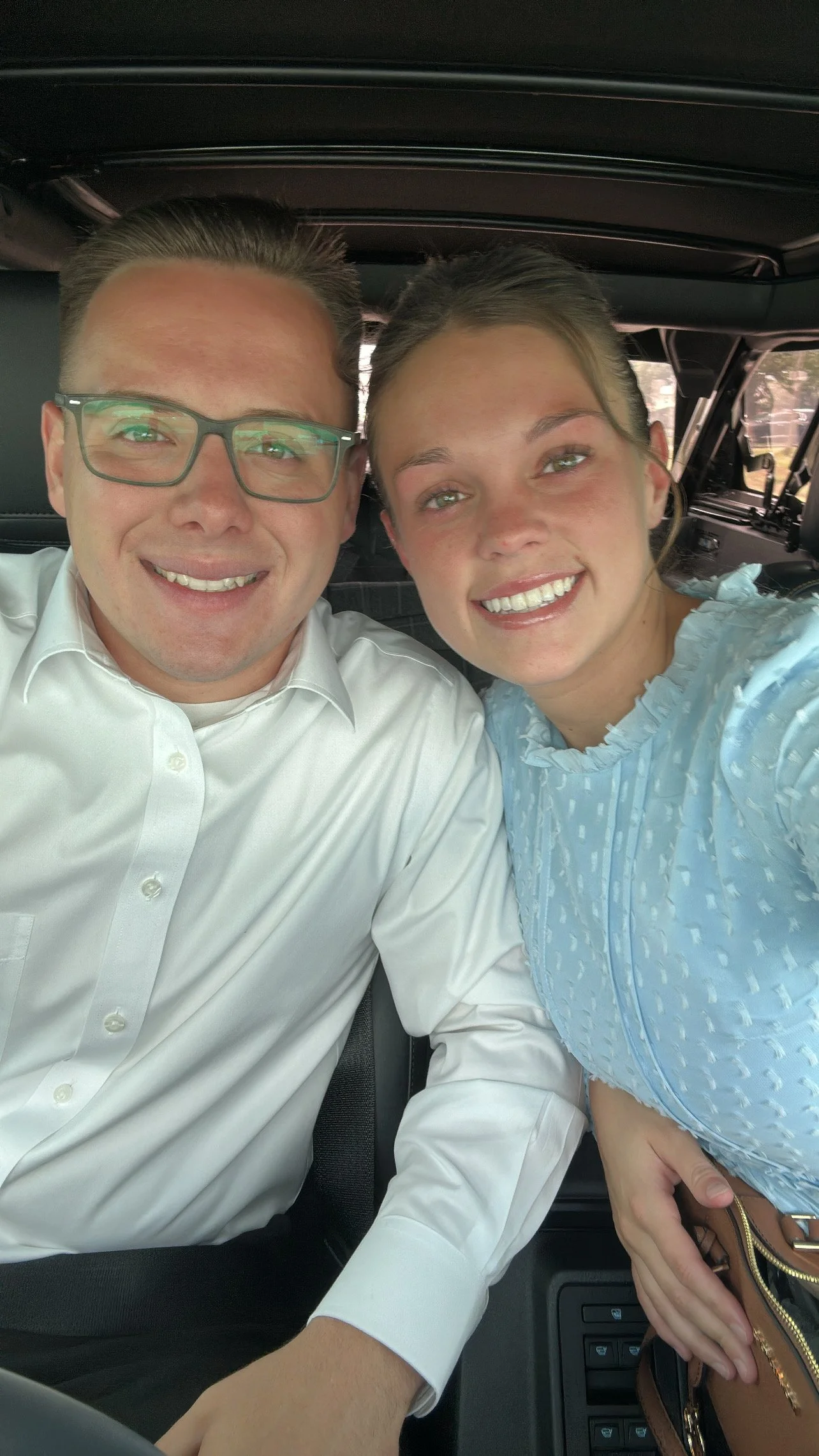 A man and woman smiling and taking a selfie inside a vehicle, with the woman wearing a light blue top and the man in a white shirt and glasses.
