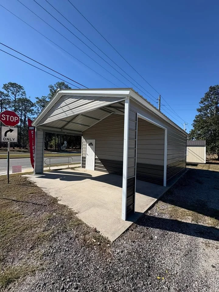 A small, metal, open-sided carport with a concrete floor, attached to a building with beige siding. Nearby are traffic signs including a STOP sign and a right turn only sign, with overhead power lines and trees in the background under a clear blue sk