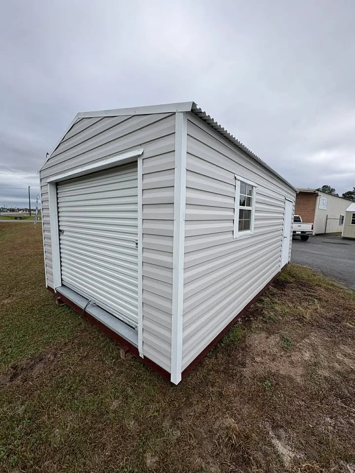 A small white metal building with a roll-up garage door, a side window, and a side door, situated on a patch of grass with a cloudy sky above.