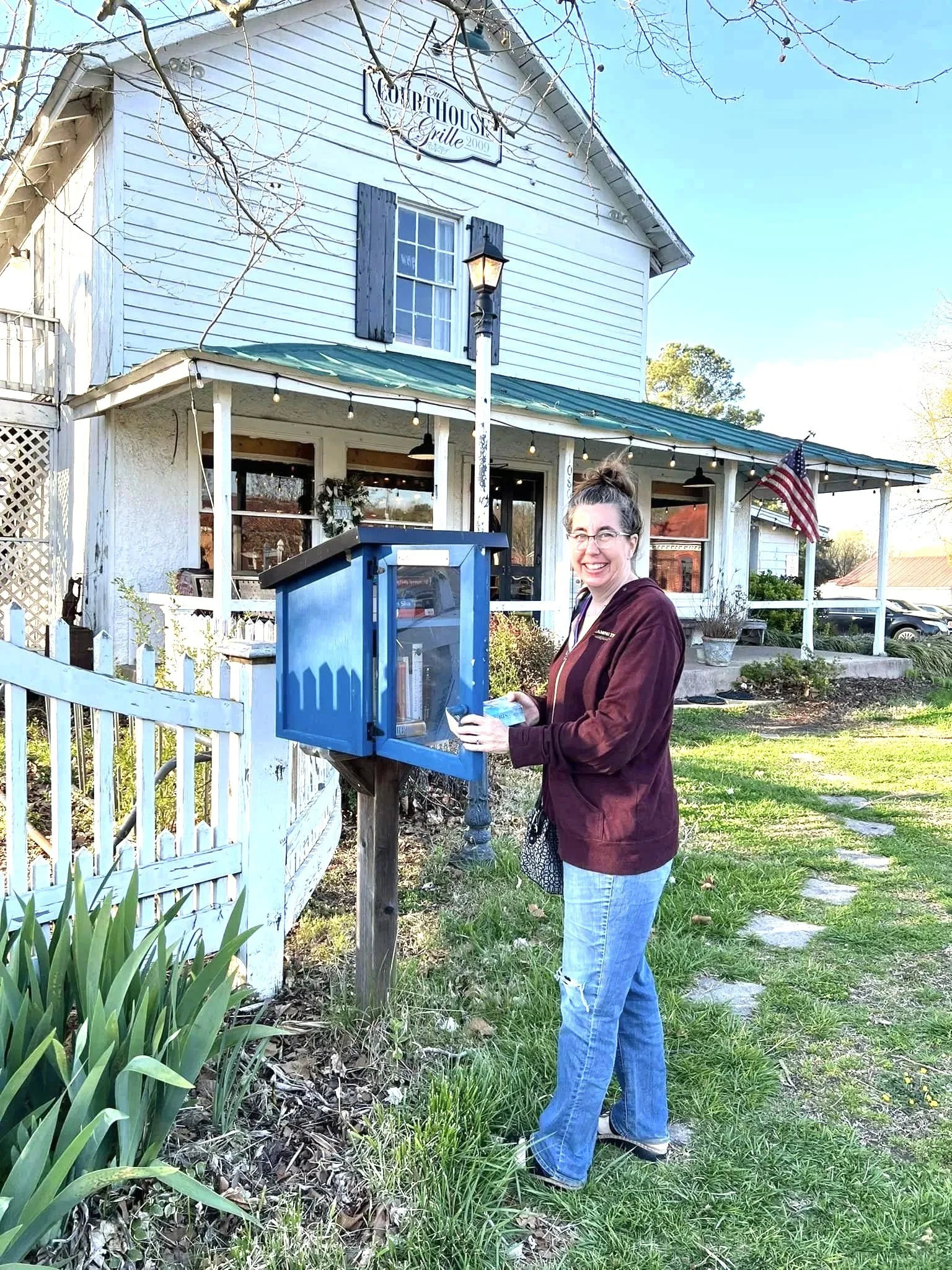 Woman standing outdoors at a blue mailbox in front of a white, two-story building with a porch and hanging string lights. The woman is smiling, holding a letter, and wearing a maroon jacket and jeans. The setting appears to be a rural or small-town area with a garden and an American flag on the porch.