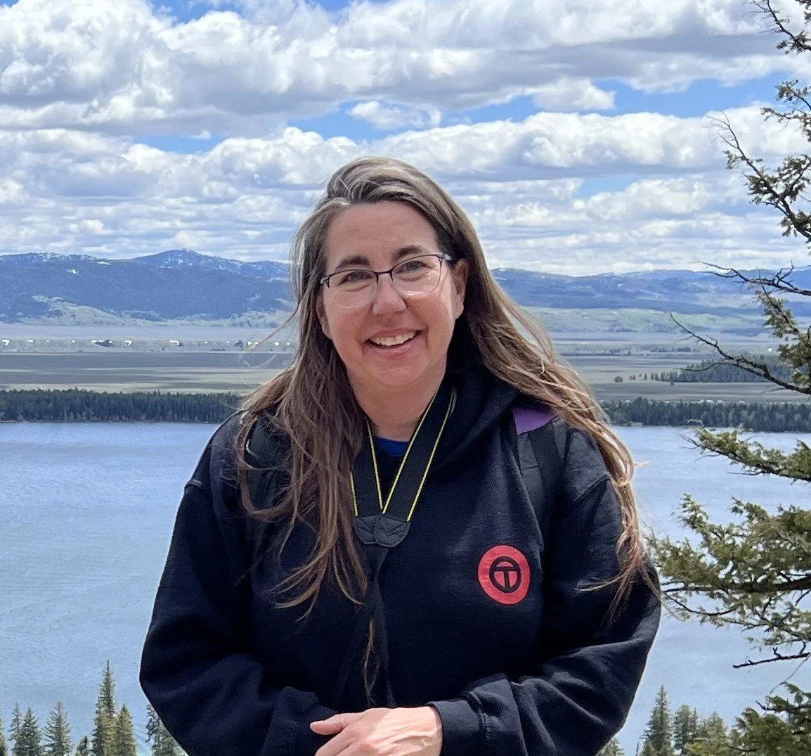 A woman with glasses smiling outdoors in front of a lake and mountains under a cloudy sky.
