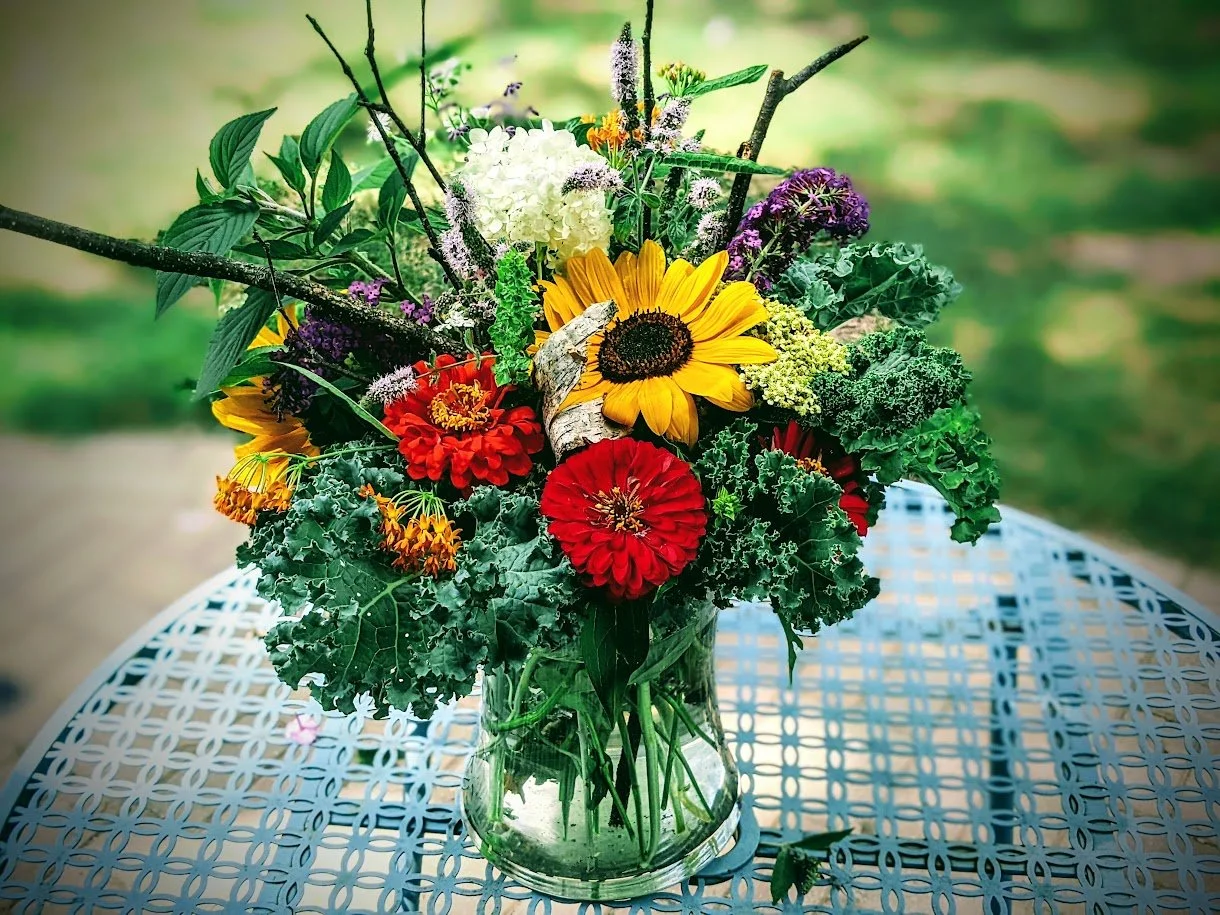 Colorful bouquet of flowers in a glass vase on a metal table outdoors, featuring sunflowers, red and orange zinnias, purple and white flowers, green leaves, and branches.