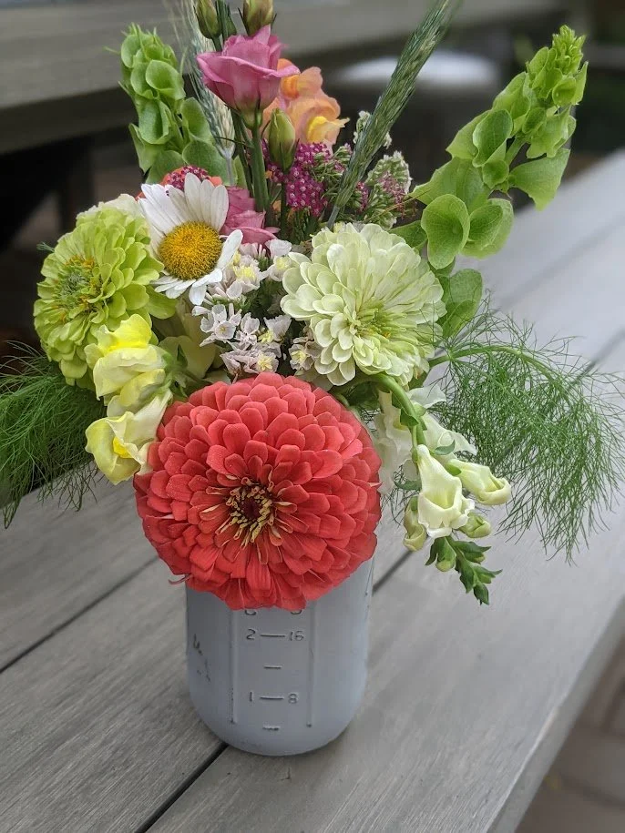 A cheerful bouquet of mixed flowers in a white measuring cup placed on a wooden table. The arrangement includes a pink ranunculus, white daisies, pale green dahlias, light yellow snapdragons, and purple, pink, and white smaller flowers with greenery.