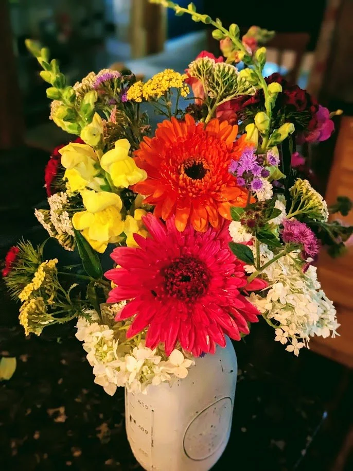 A colorful bouquet of various flowers including red and orange gerbera daisies, yellow snapdragons, purple and white statice, and green foliage in a white vase.