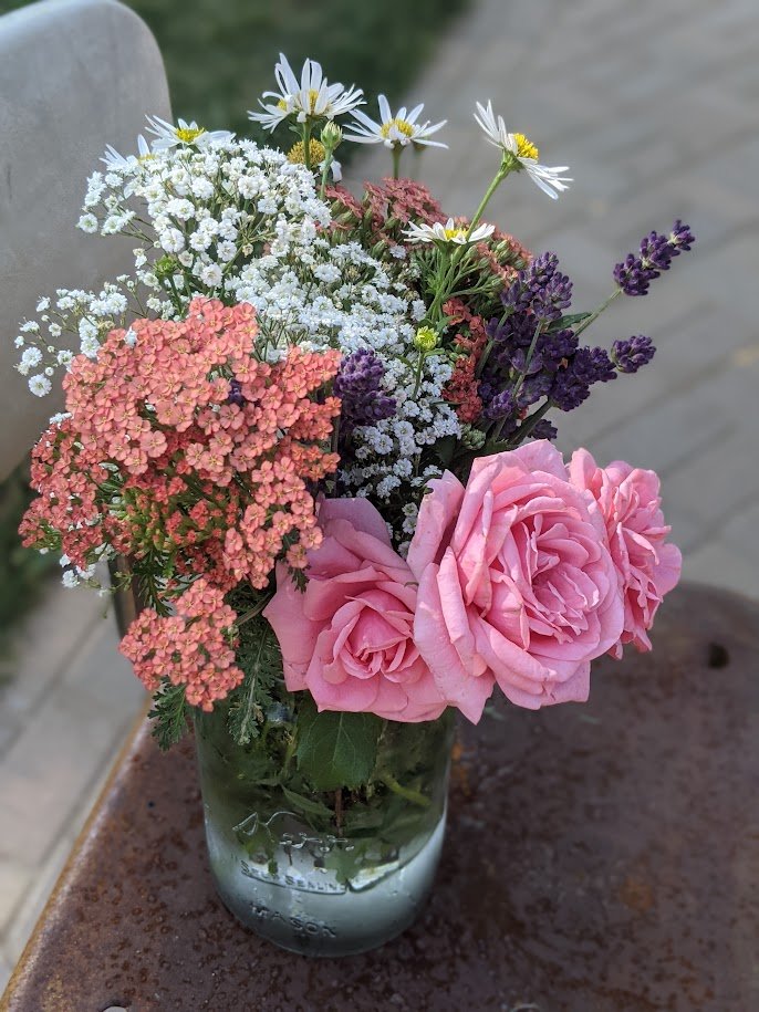 A glass jar filled with a colorful assortment of flowers, including pink roses, white daisies, purple lavender, and small white and pink blooms, placed on a rusty surface outdoors.