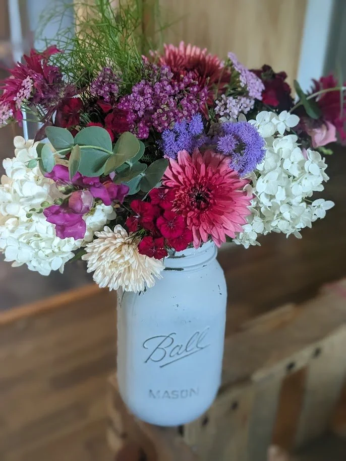 A bouquet of mixed flowers in a white Mason jar with the word 'Ball' written on it. The bouquet includes hydrangeas, dahlias, sweet peas, alliums, and various greens.