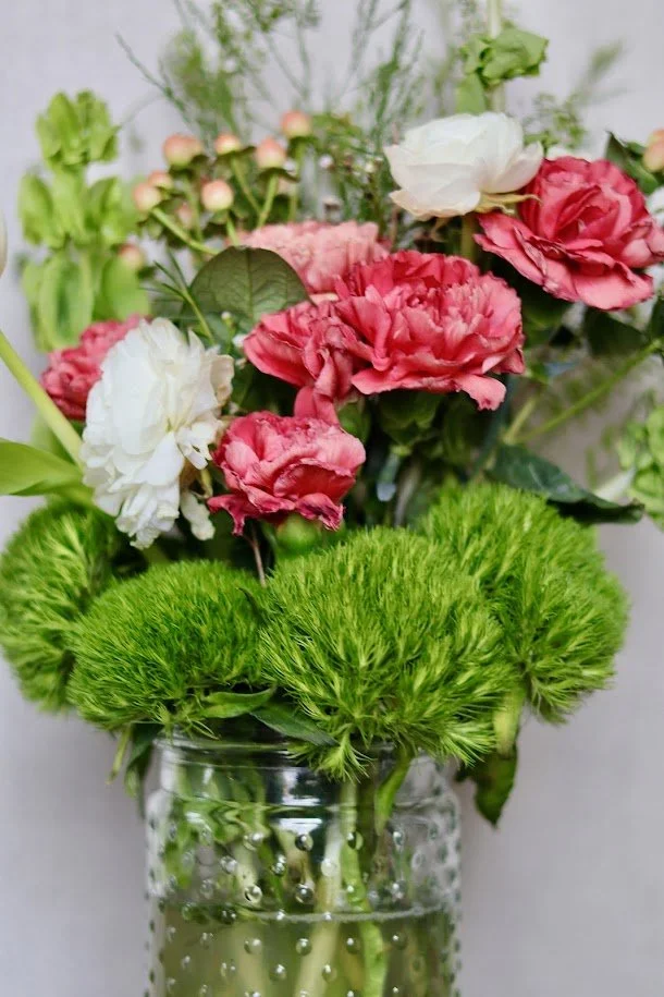 A glass jar filled with water holds a bouquet of pink, white, and green flowers, including lush green blooms, pink and white carnations, and small pink buds, arranged with greenery.