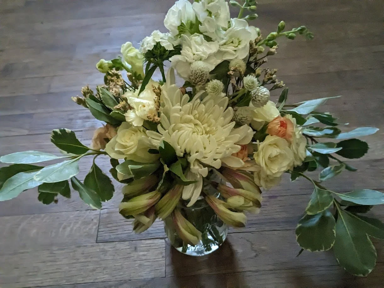 A bouquet of white and cream flowers with green leaves in a glass vase, placed on a wooden floor.