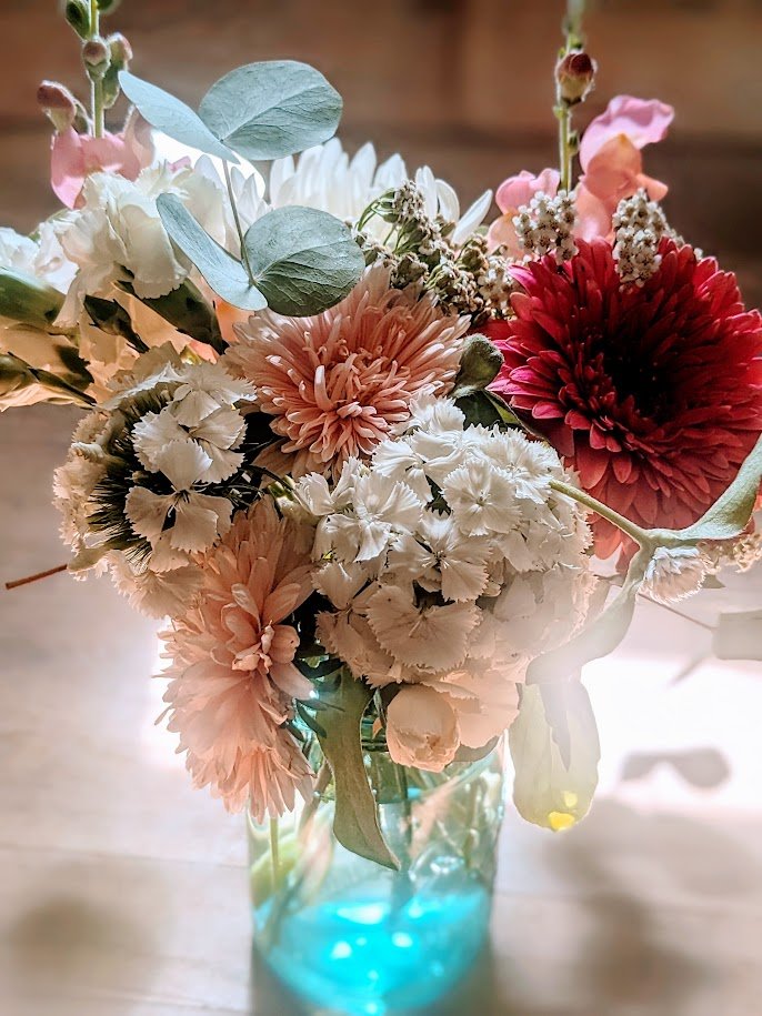 A colorful bouquet of flowers in a blue glass vase, including pink, white, peach, and red blooms, with hints of green leaves and stems.