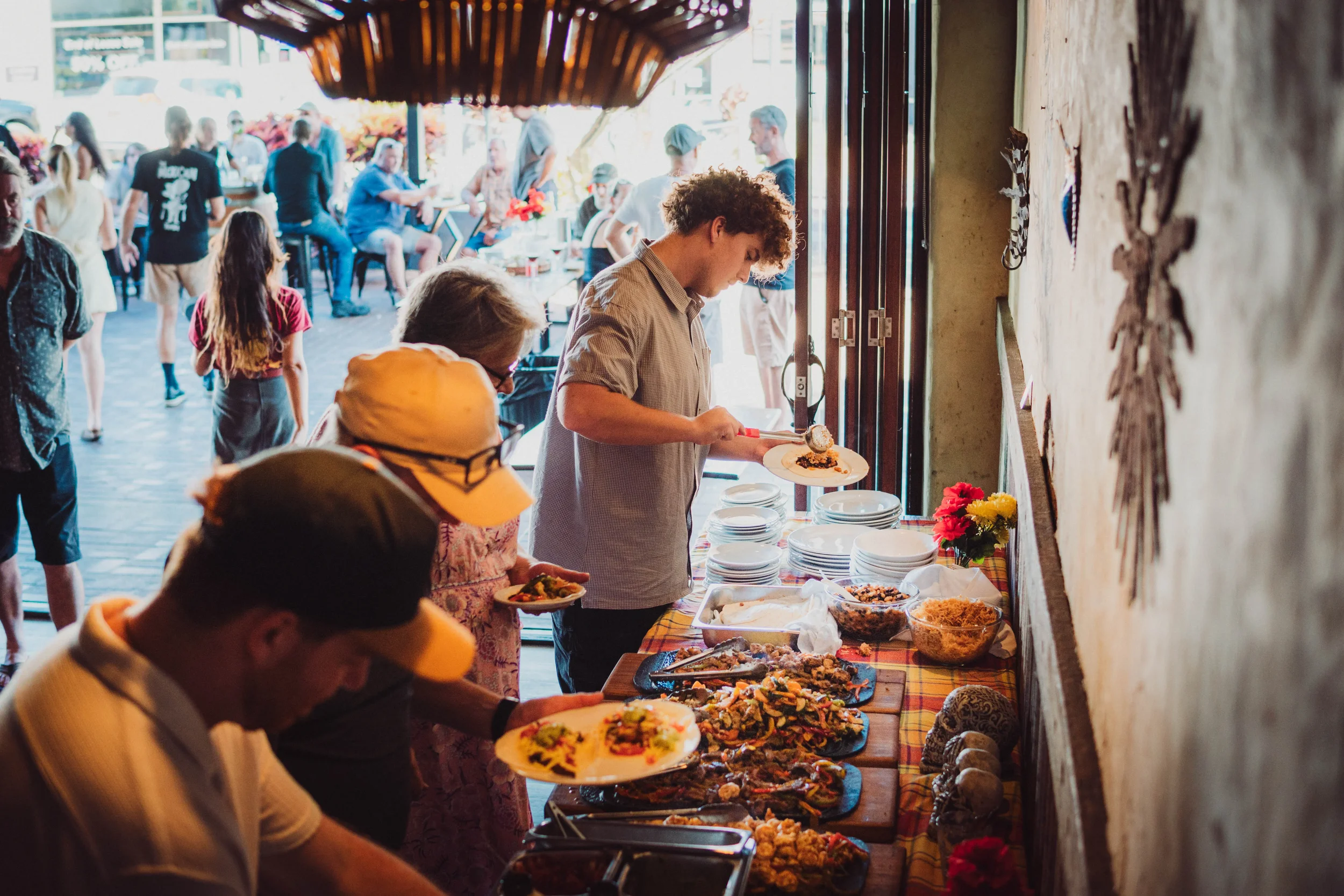 People serving themselves food from a buffet at a social gathering or party, with several dishes on a table near a window, and more people in the background outside.