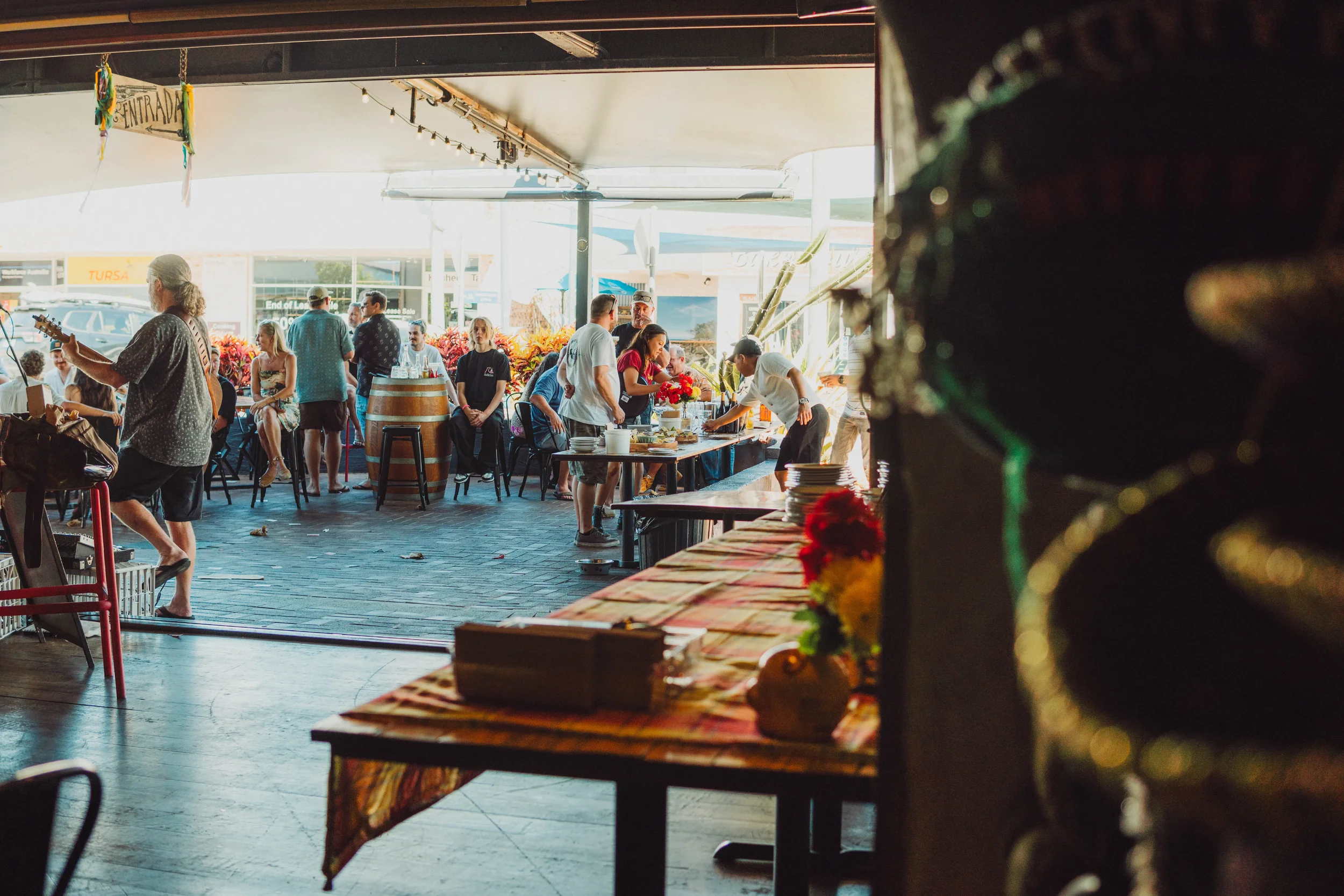 People dining and socializing at a restaurant patio with tables, chairs, and decorations, seen from inside the restaurant.