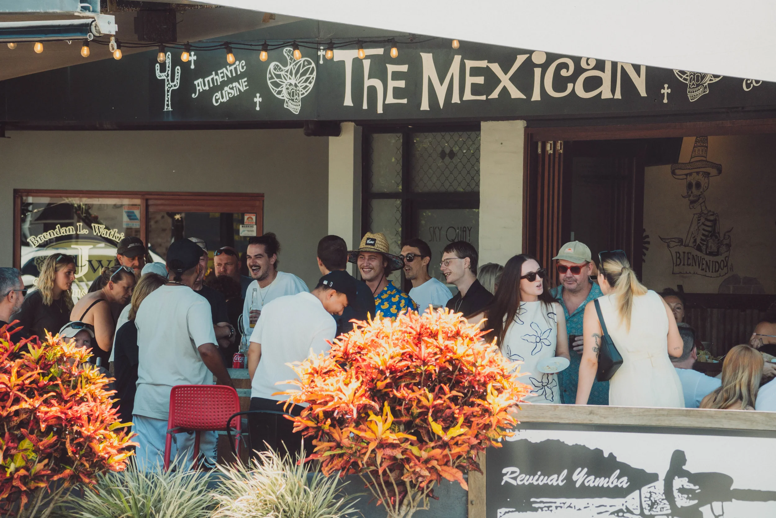 People gathered outside a restaurant called 'The Mexican' with a sign reading 'Authentic Cuisine' and art featuring skeletons, including a skeleton wearing a sombrero.