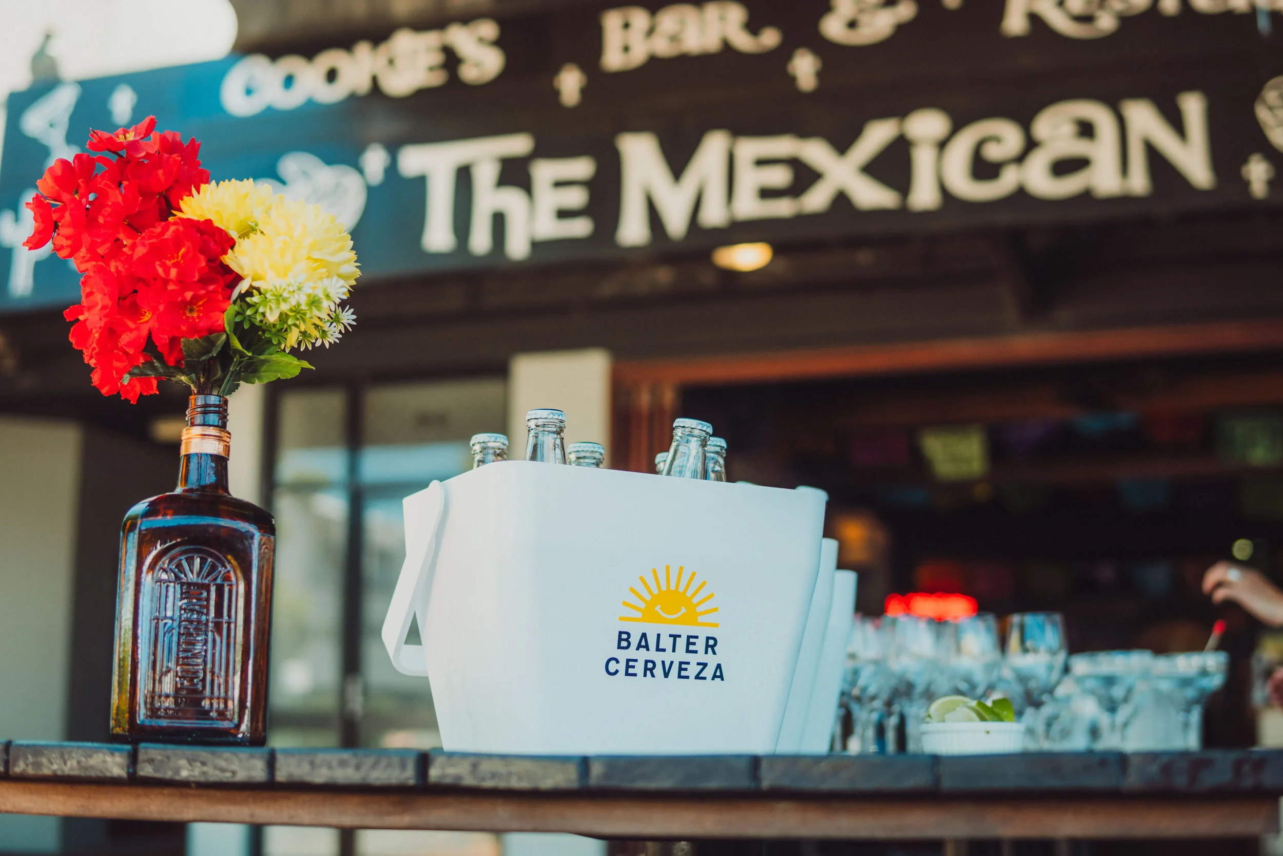 A table with a brown beer bottle holding red, yellow, and white flowers, and a white bucket with bottled drinks labeled 'BALTER CERVEZA.' In the background, a sign says 'Cookies + The Mexican'.