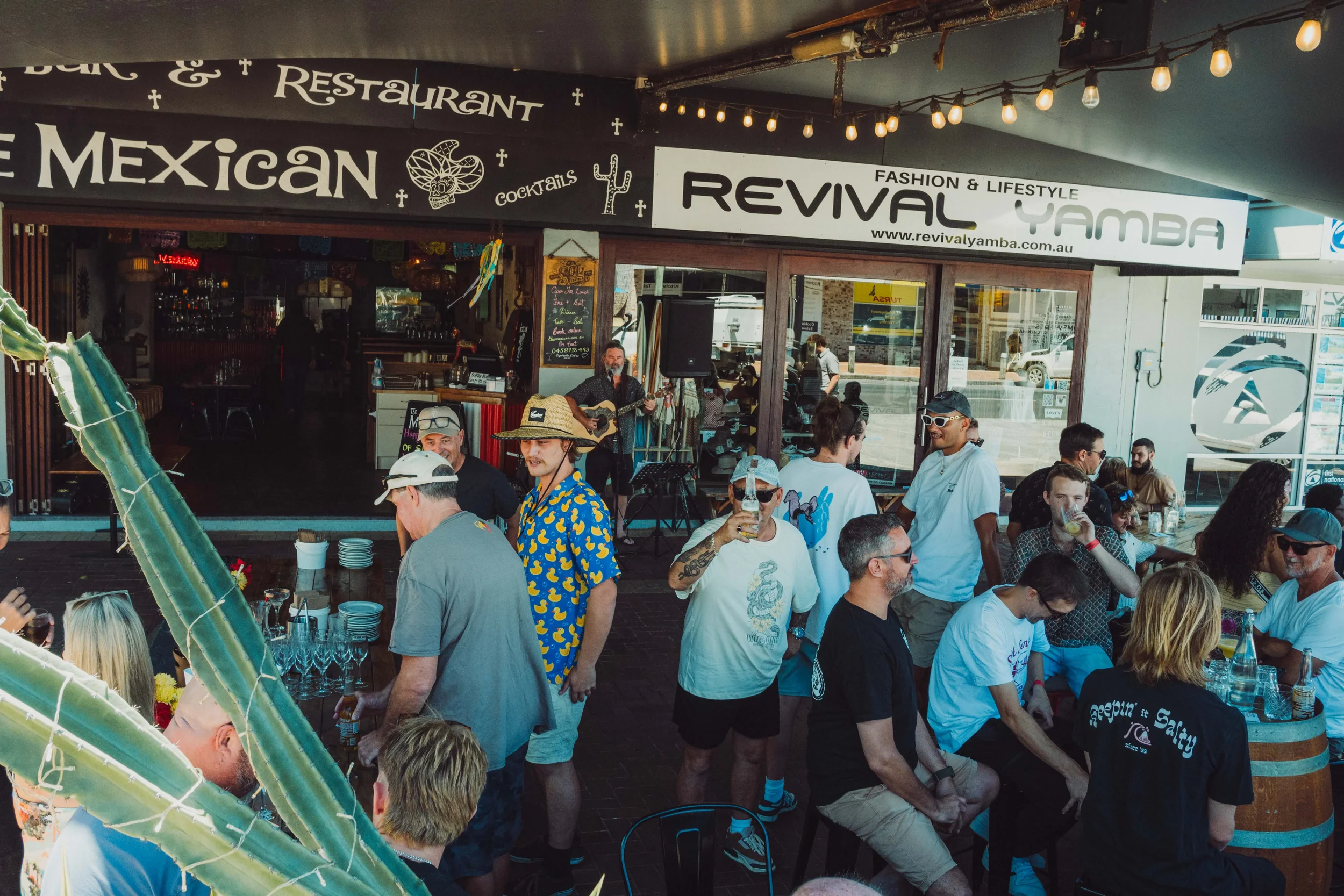 Outdoor gathering at a Mexican restaurant with live music, many people enjoying drinks, some standing and some seated, in front of a restaurant called Revival Yamba.