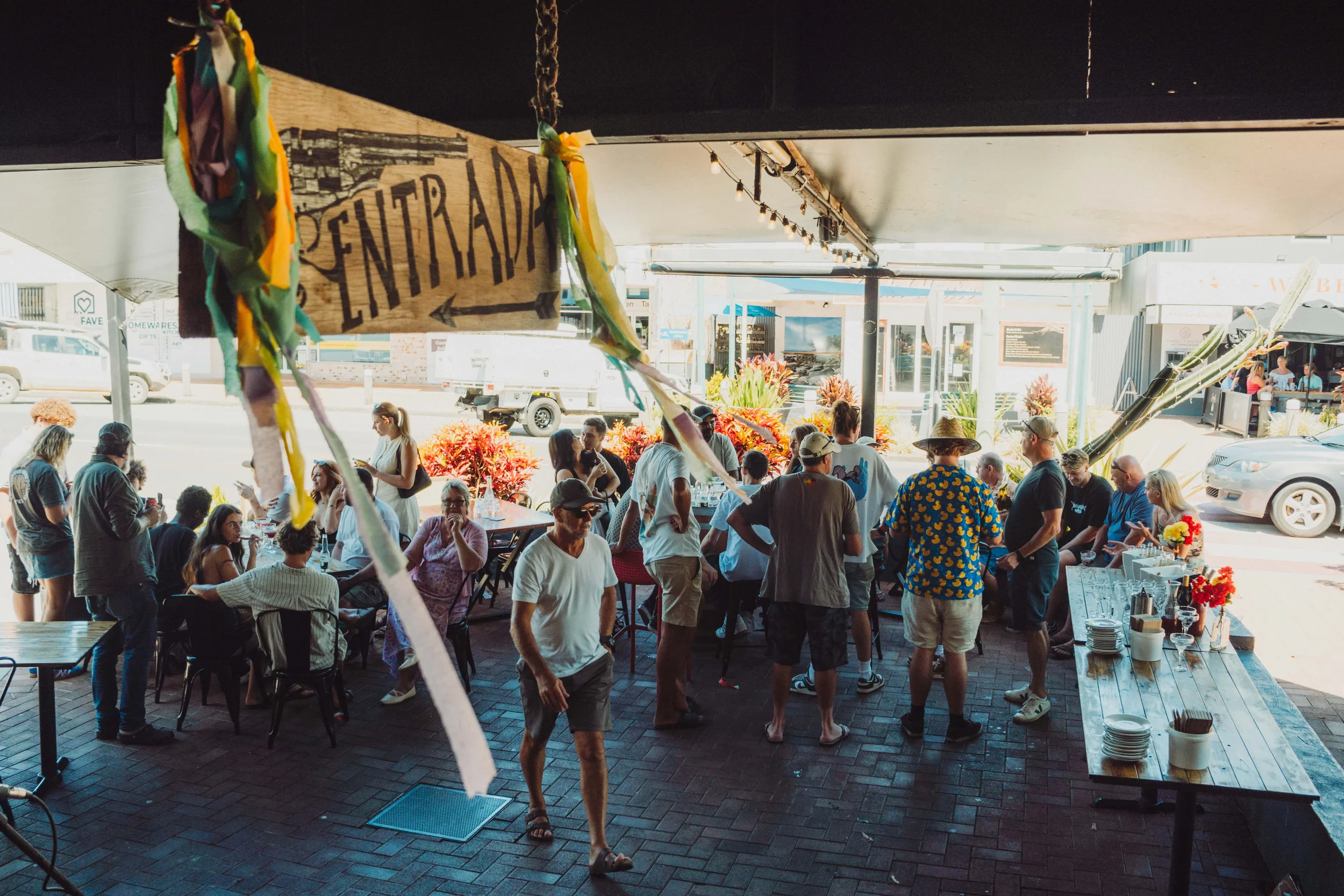 A lively outdoor dining scene with people gathered under a shaded area, some seated and some standing, and a sign pointing to the entrance.