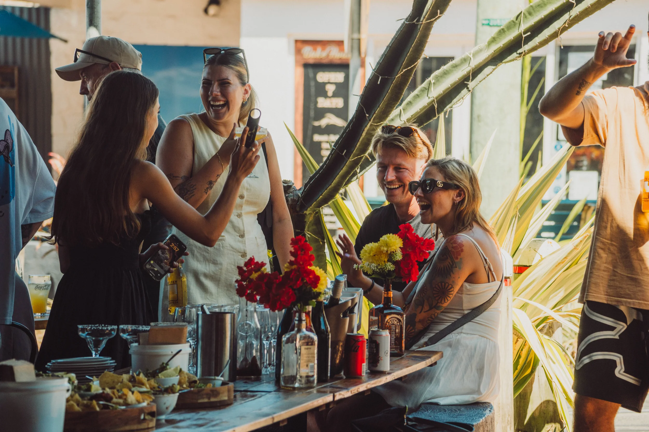 Group of friends enjoying a social gathering at a bar or restaurant, laughing and taking pictures, with tables of drinks and snacks, surrounded by tropical plants.