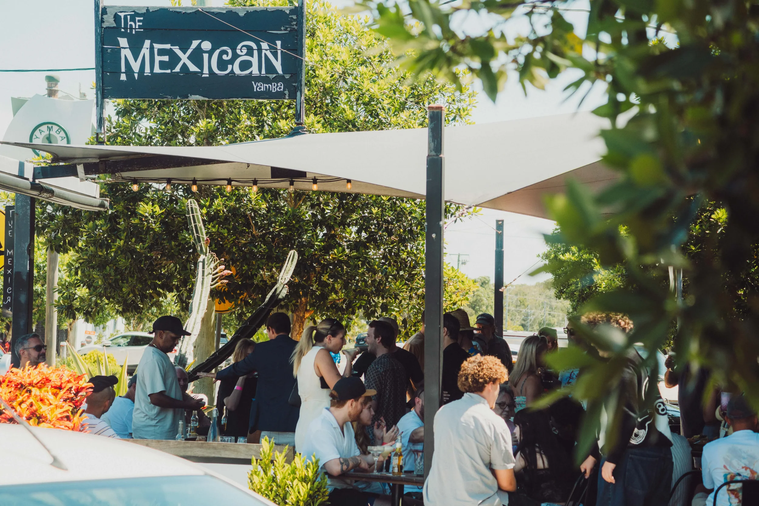 People gather at an outdoor restaurant or bar with a sign that says 'The Mexican Yamba'; some are sitting at tables, others are standing, and there are large trees and potted plants around the seating area. The scene appears lively and social on a su