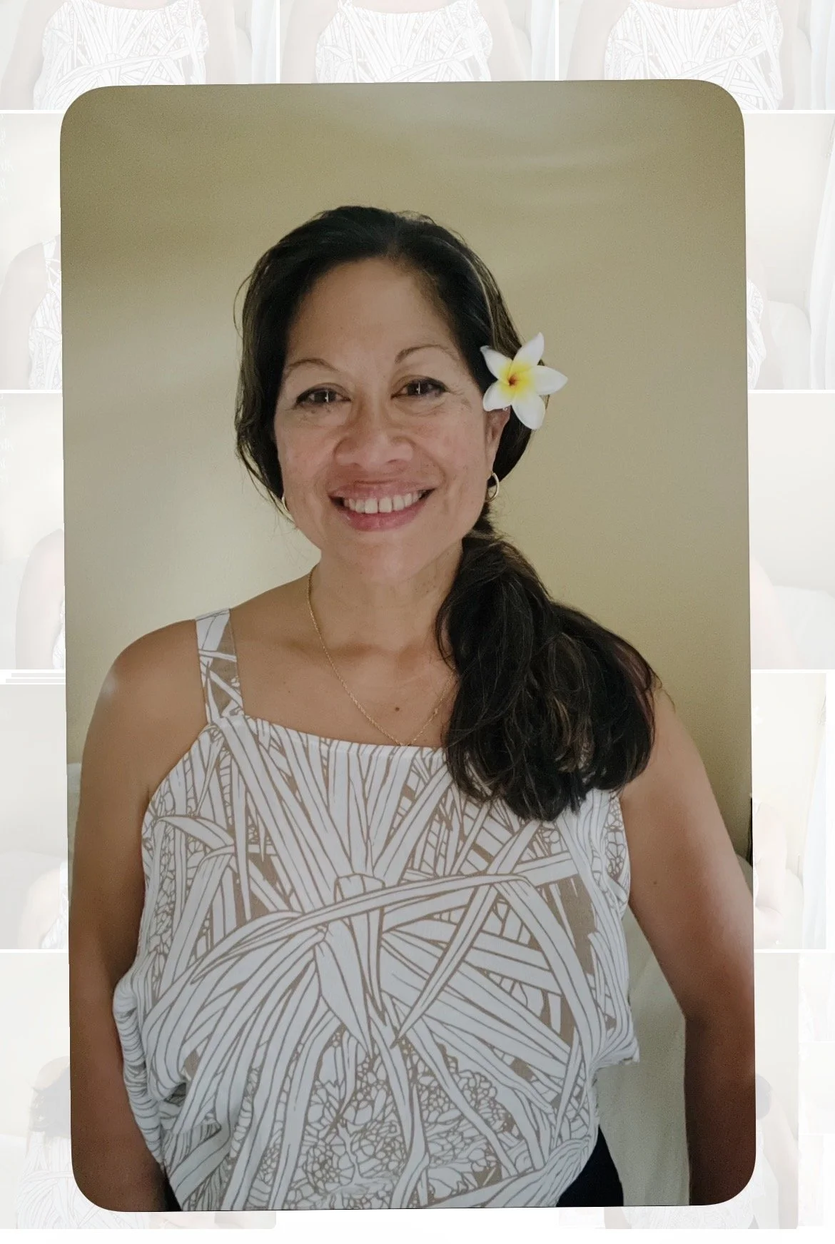 Smiling woman with dark hair, a white flower tucked behind her ear, wearing a patterned sleeveless top, standing against a plain light-colored wall.