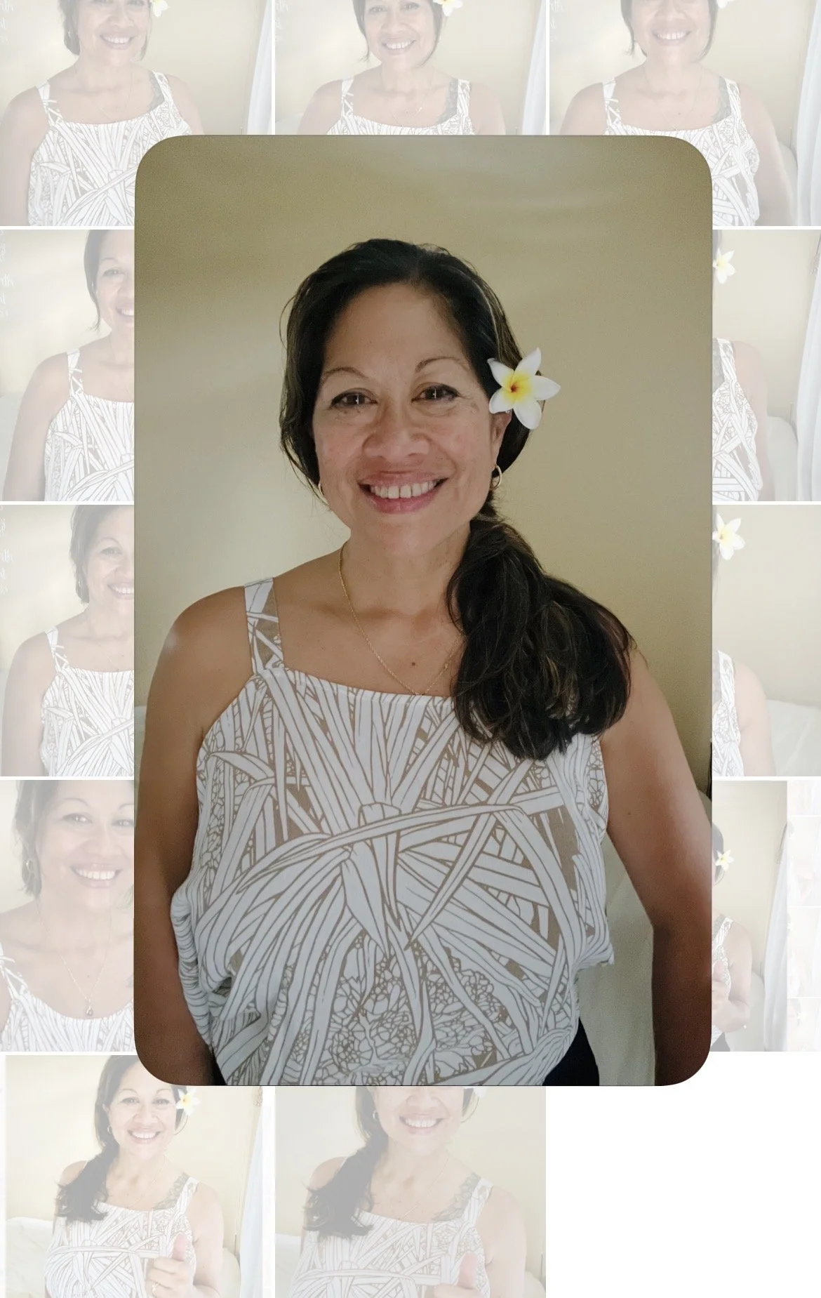 A woman with dark hair and a white flower in her hair, smiling, wearing a white and brown patterned top, standing against a beige wall.