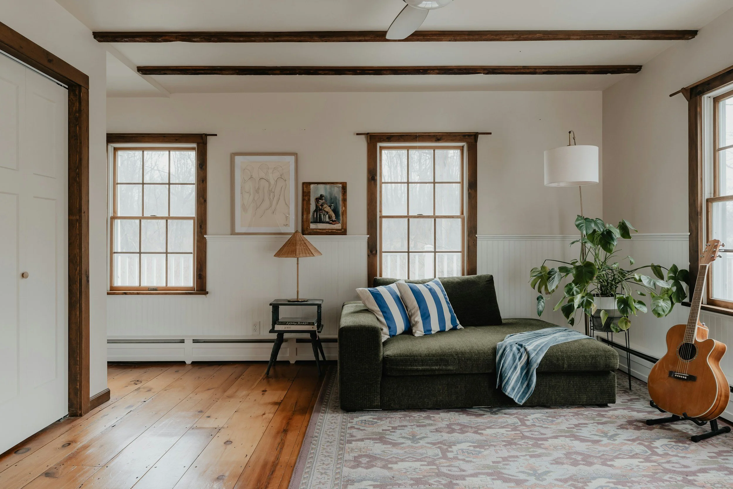 Living room with wooden floor and white walls, featuring two windows with wooden trim, a dark green sofa with striped pillows, a potted plant, a guitar, a side table with a lamp, and framed artwork on the wall.