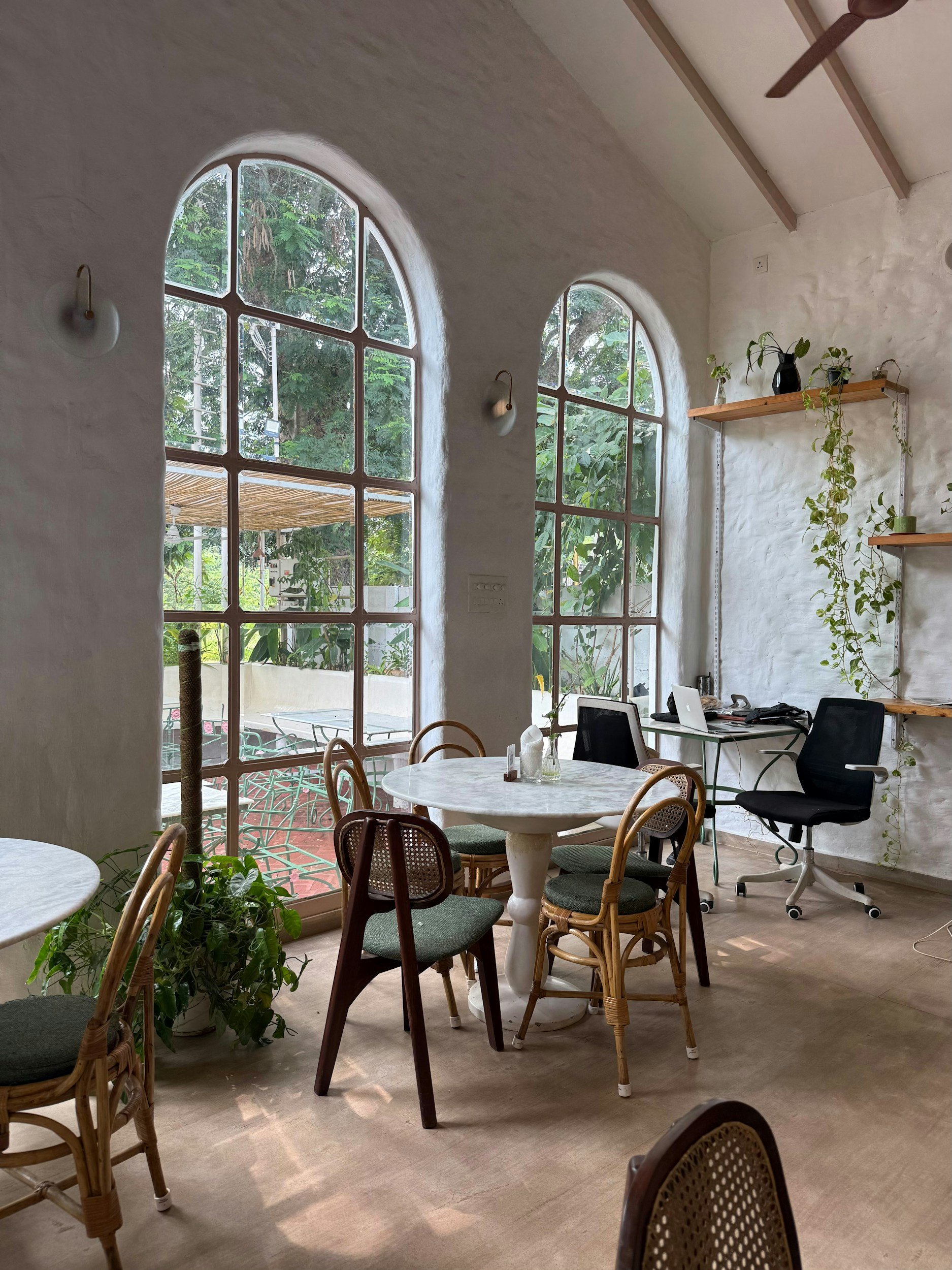 Interior of a cozy cafe or workspace with large arched windows, white textured walls, and wooden accents. Various chairs surround a round marble table, and a desk with a laptop and office chair are visible in the corner. Shelves with plants are on the wall, adding greenery.