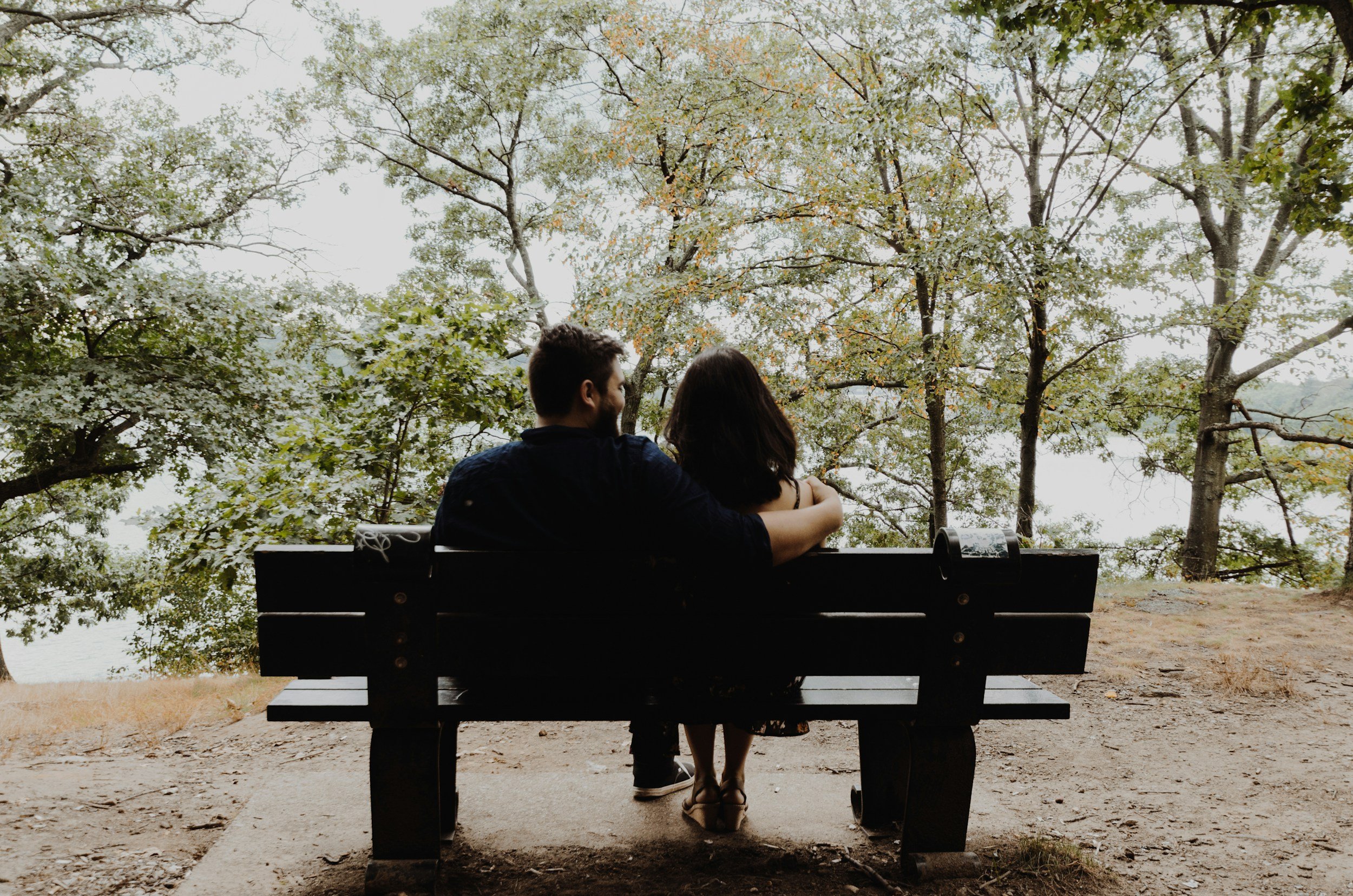 A young couple sitting on a park bench, embracing and looking at each other, surrounded by trees with green leaves.