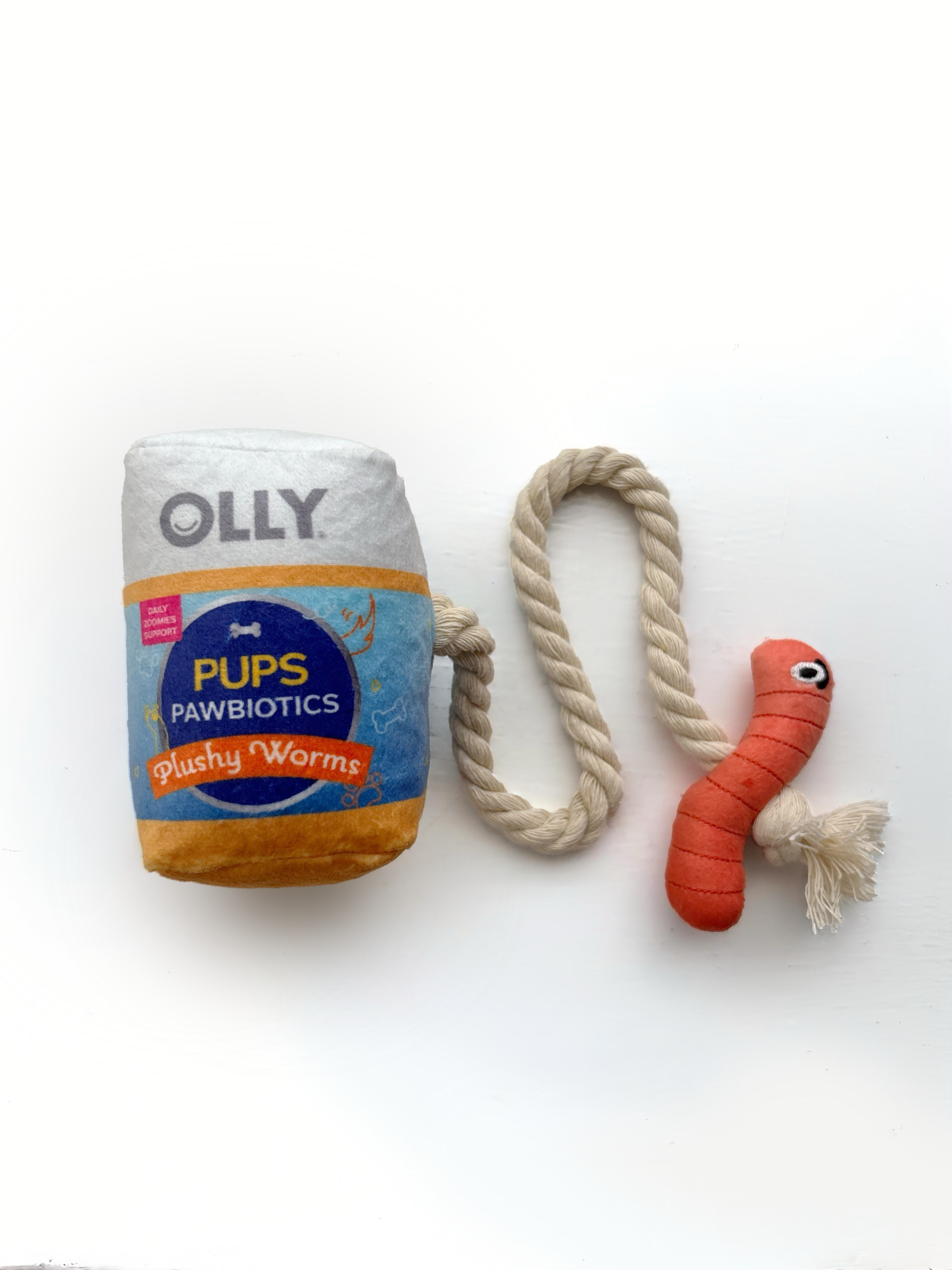 A dog toy reading "Pups Pawbiotic Plushy Worms" and resembling an OLLY bottle connected to a beige rope and an orange plush worm toy with embroidered eyes on a white background.