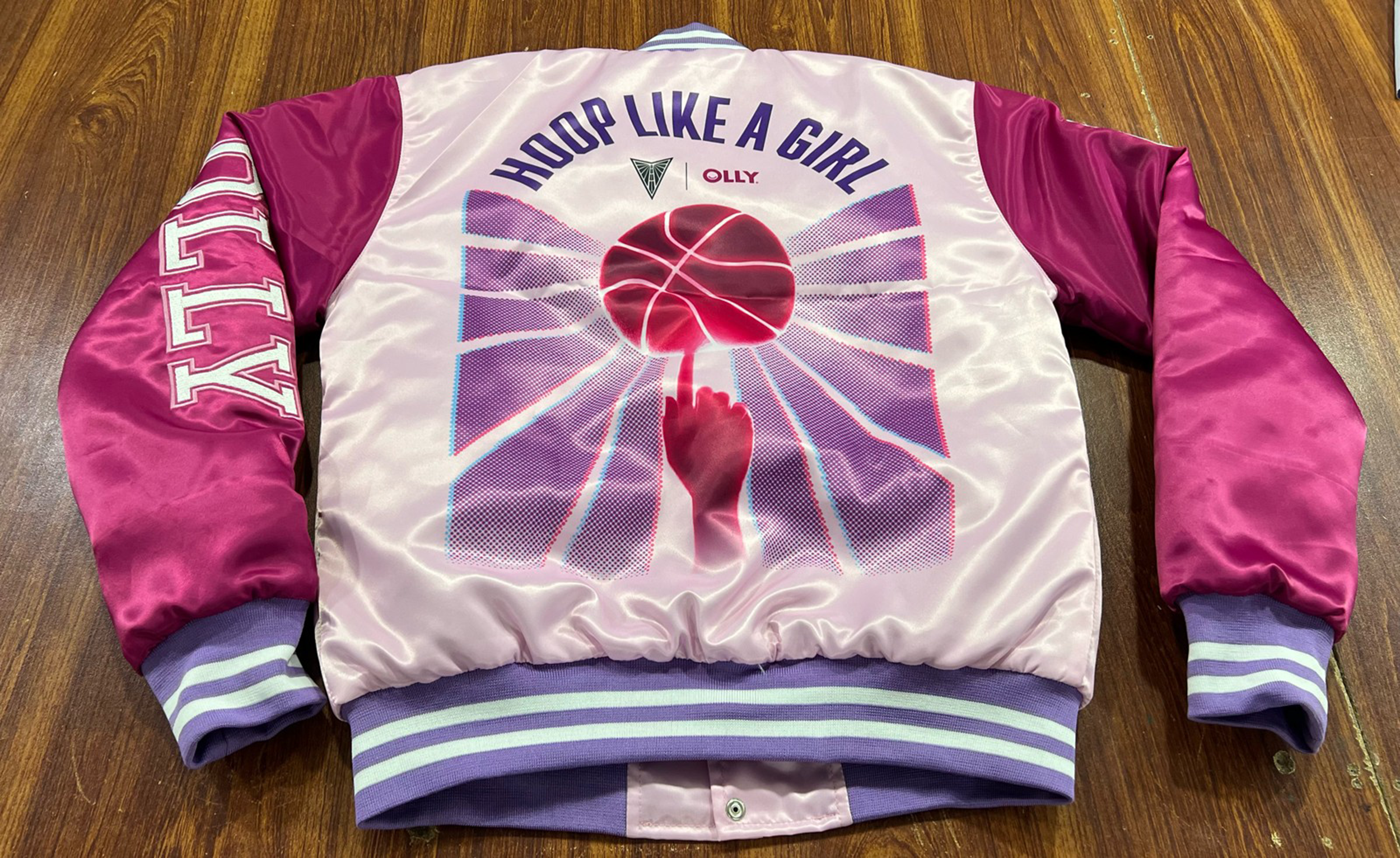 A satin bomber jacket with a colorful graphic on the back featuring a finger balancing a basketball and the words 'HOOP LIKE A GIRL' in purple with the Valkyries and OLLY logos.