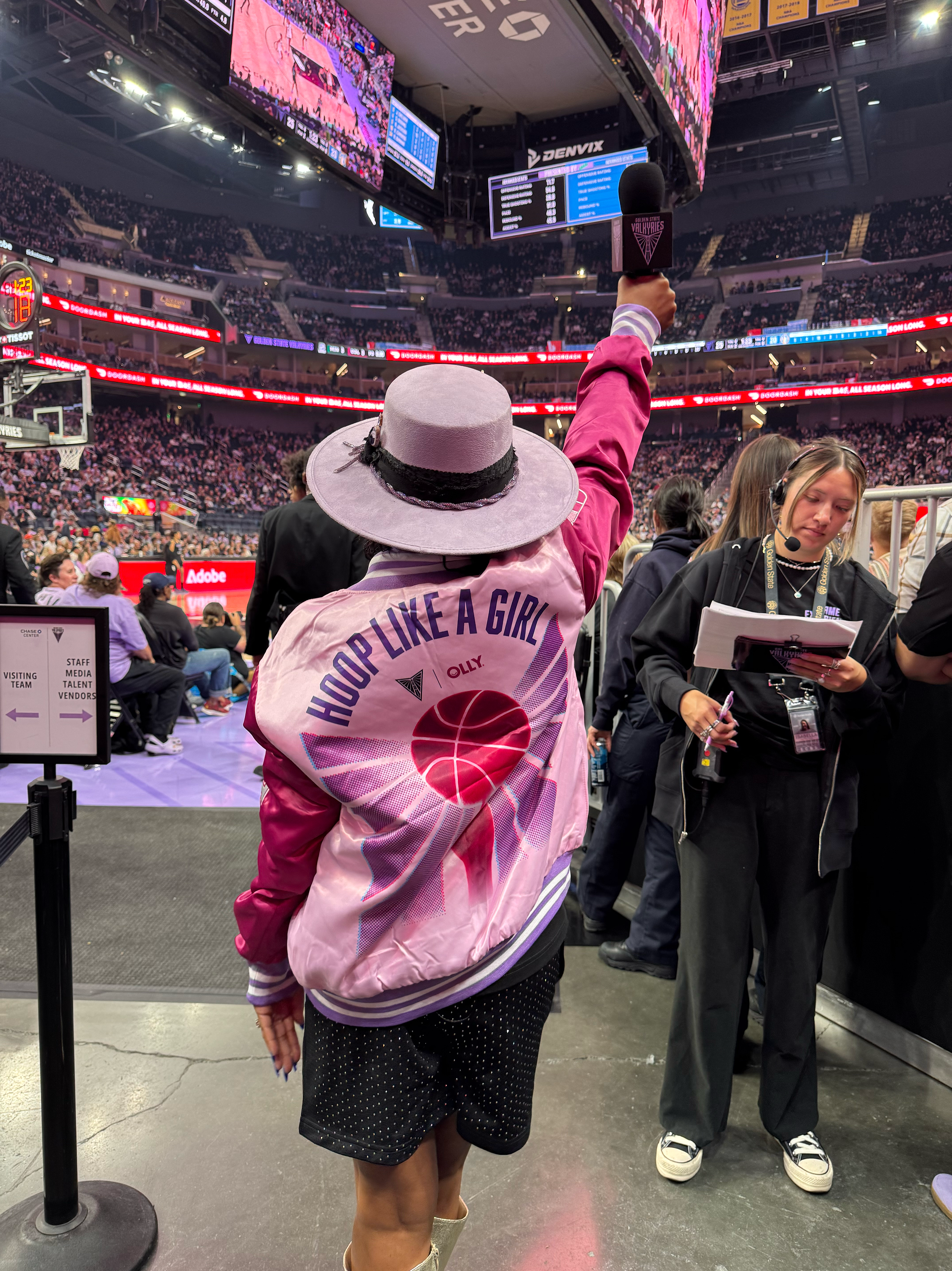 A person wearing a pink jacket with a basketball graphic and the words 'HOP LIKE A GIRL' on the back, holding up a microphone at a Valkyries basketball game. The arena is filled with spectators, large screens, and electronic scoreboards overhead.