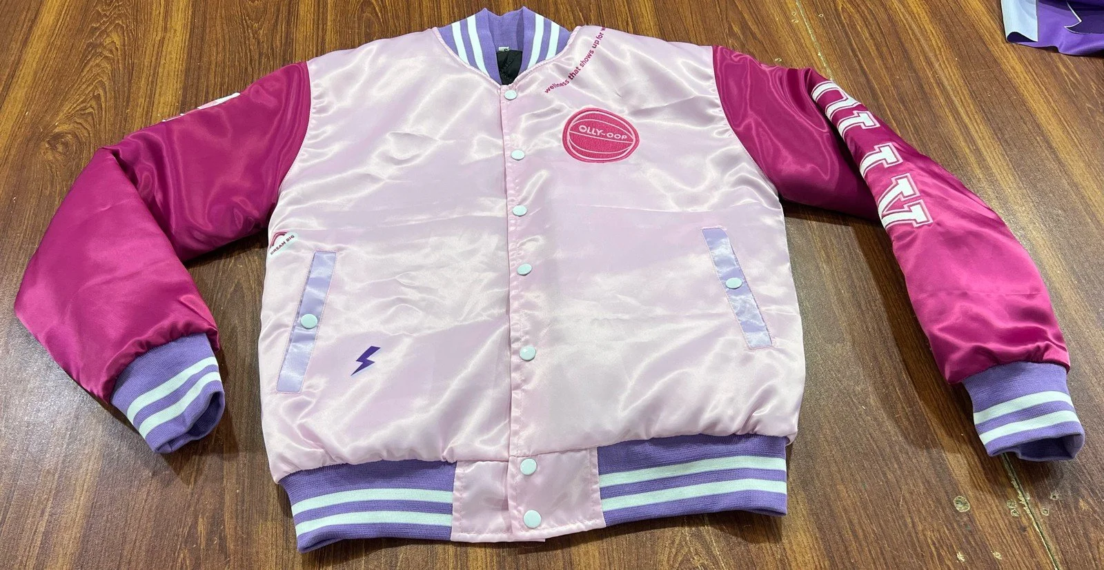 Pink satin varsity jacket with magenta sleeves; lavender striped collar, cuffs, and waistband, featuring patches and embroidery on the front and sleeves reading "OLLY."