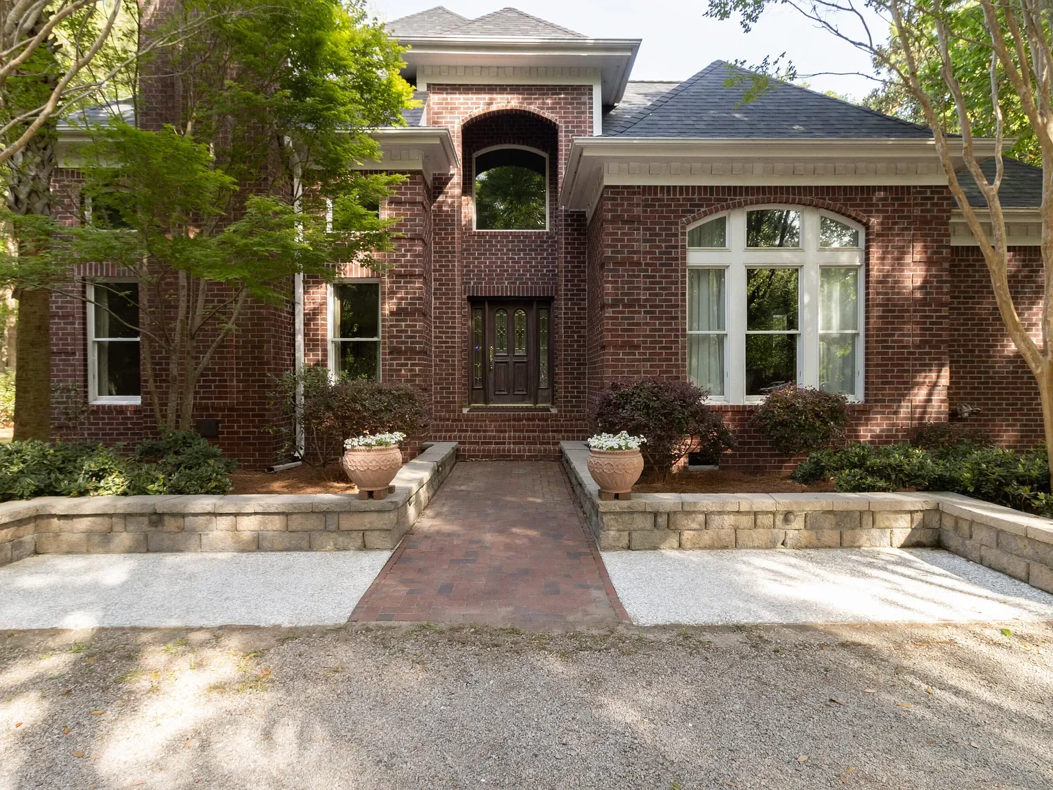 Front view of a brick house with large windows, surrounded by trees and a landscaped yard with potted plants, brick walkway, and a stone border.