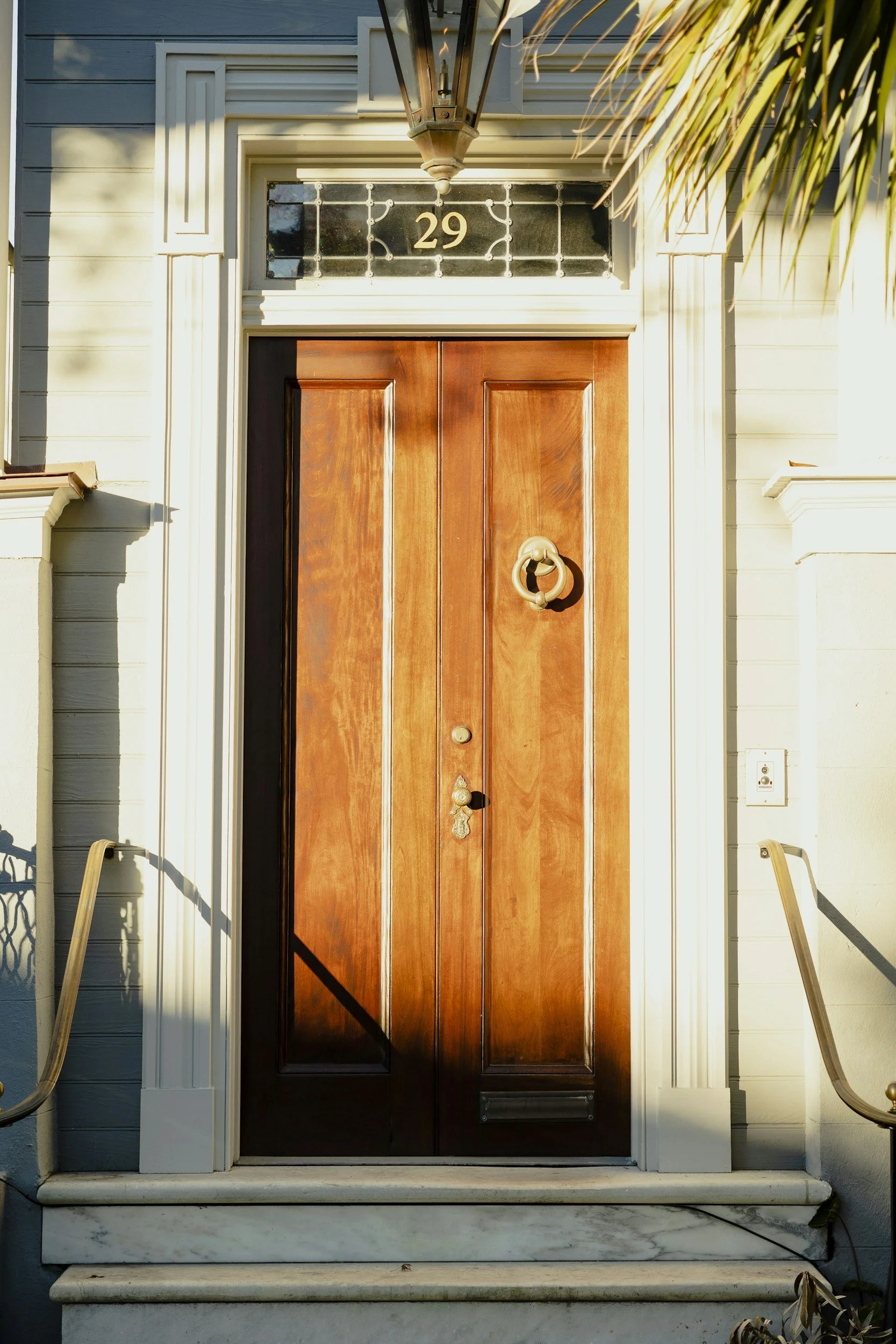 Front door of a house made of polished wood with a metal door knocker, a doorbell, and a peephole. The house number 29 above the door, decorative trim surrounding the door, and white steps leading up to it. There are shadows and sunlight on the house.