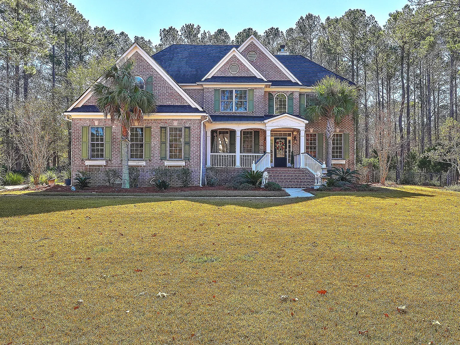 A large two-story brick house with green shutters, surrounded by trees and a well-manicured lawn. The house features a front porch with white railings and steps leading up to the front door, which is decorated with a wreath. There are palm trees in t