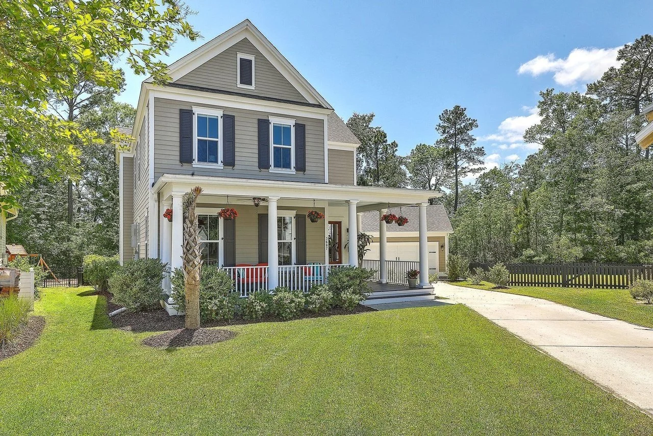 A two-story house with gray siding, white trim, and black shutters, featuring a front porch with four white columns and hanging flower baskets, surrounded by green lawn and trees under a blue sky with scattered clouds.