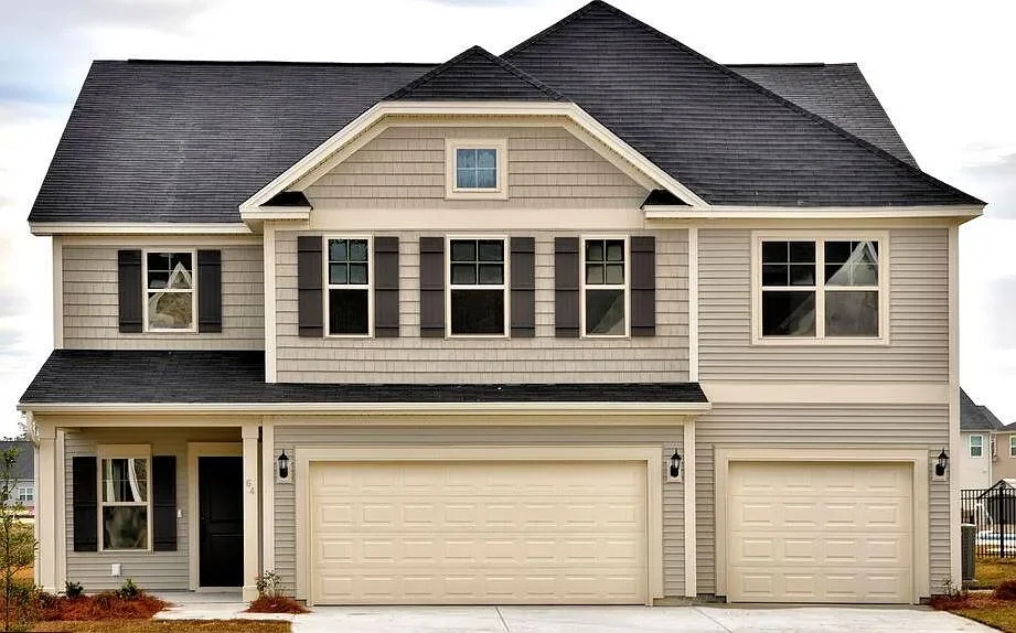 A two-story suburban house with beige siding and black shutters, featuring a double garage, a small front porch, and multiple windows.