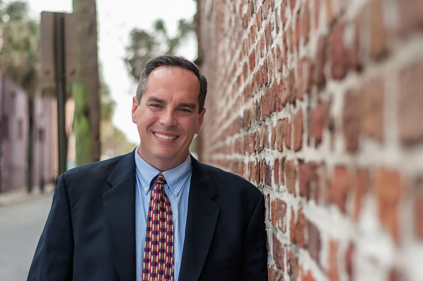 A smiling man in a blue dress shirt, colorful patterned tie, and dark blazer leaning against a brick wall outdoors.