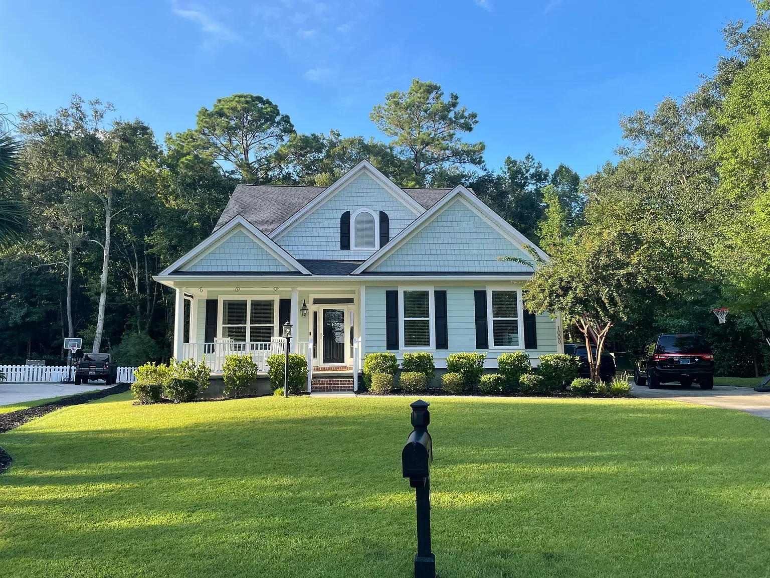 A white, two-story house with black shutters and a small front porch, surrounded by green lawn and trees, under a blue sky.