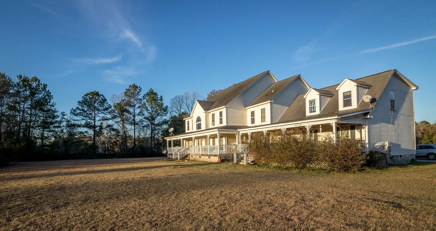 A large two-story white house with a wrap-around porch, multiple gabled roofs, and satellite dishes in a spacious yard with trees in the background under a blue sky.