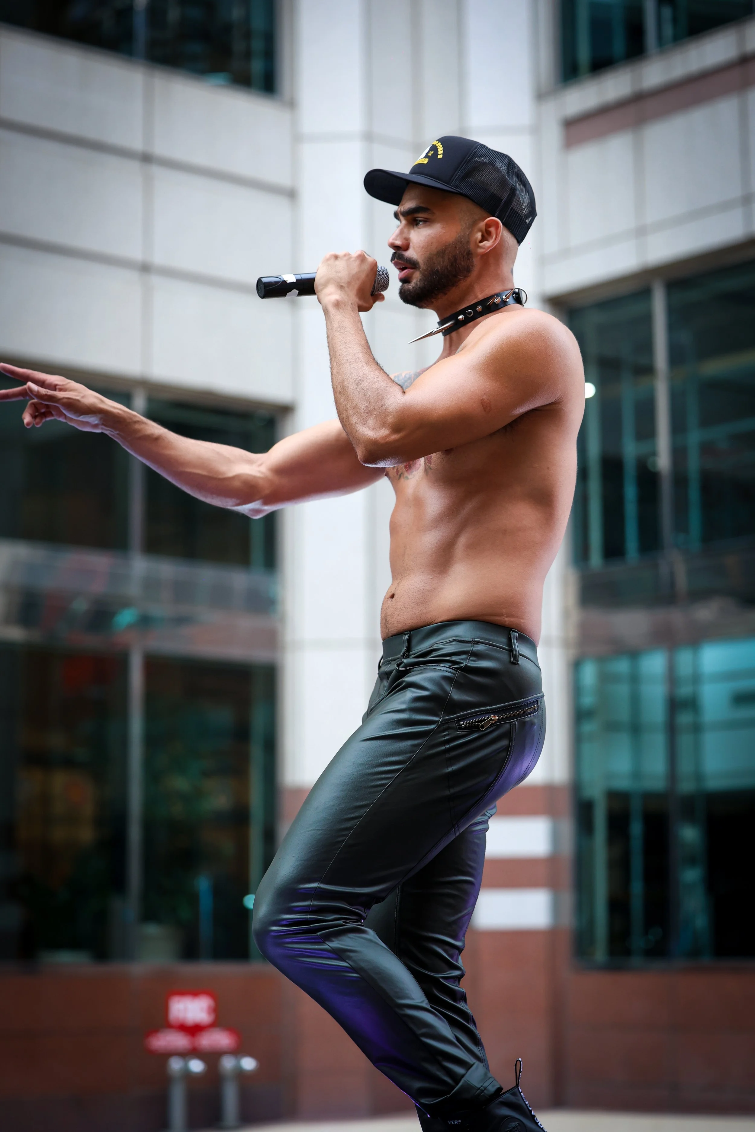 A shirtless man with a beard, wearing a black trucker cap, leather pants, and a spiked choker, performing outdoors with a microphone in hand.