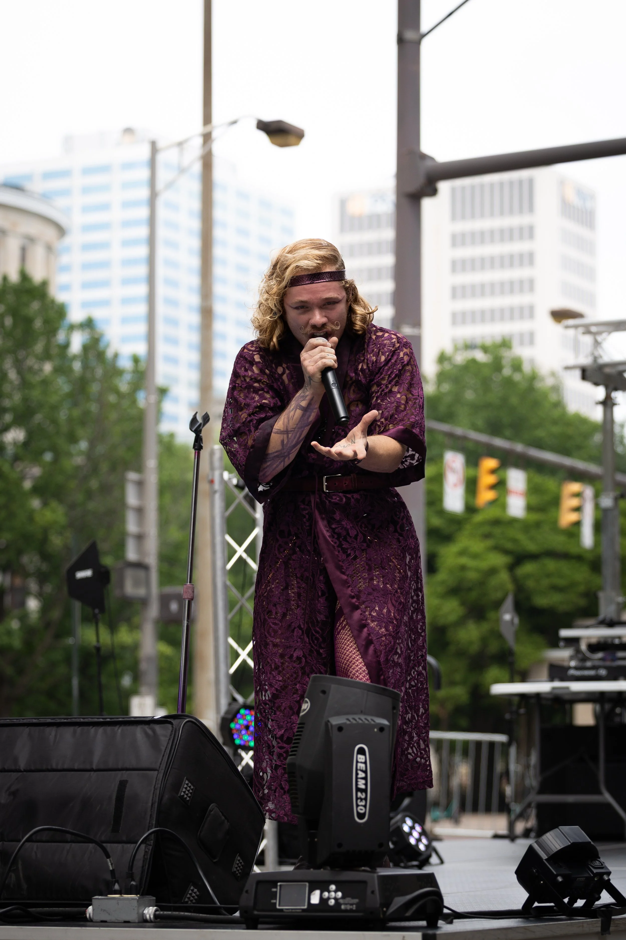 Performer singing on an outdoor stage with city buildings in the background.