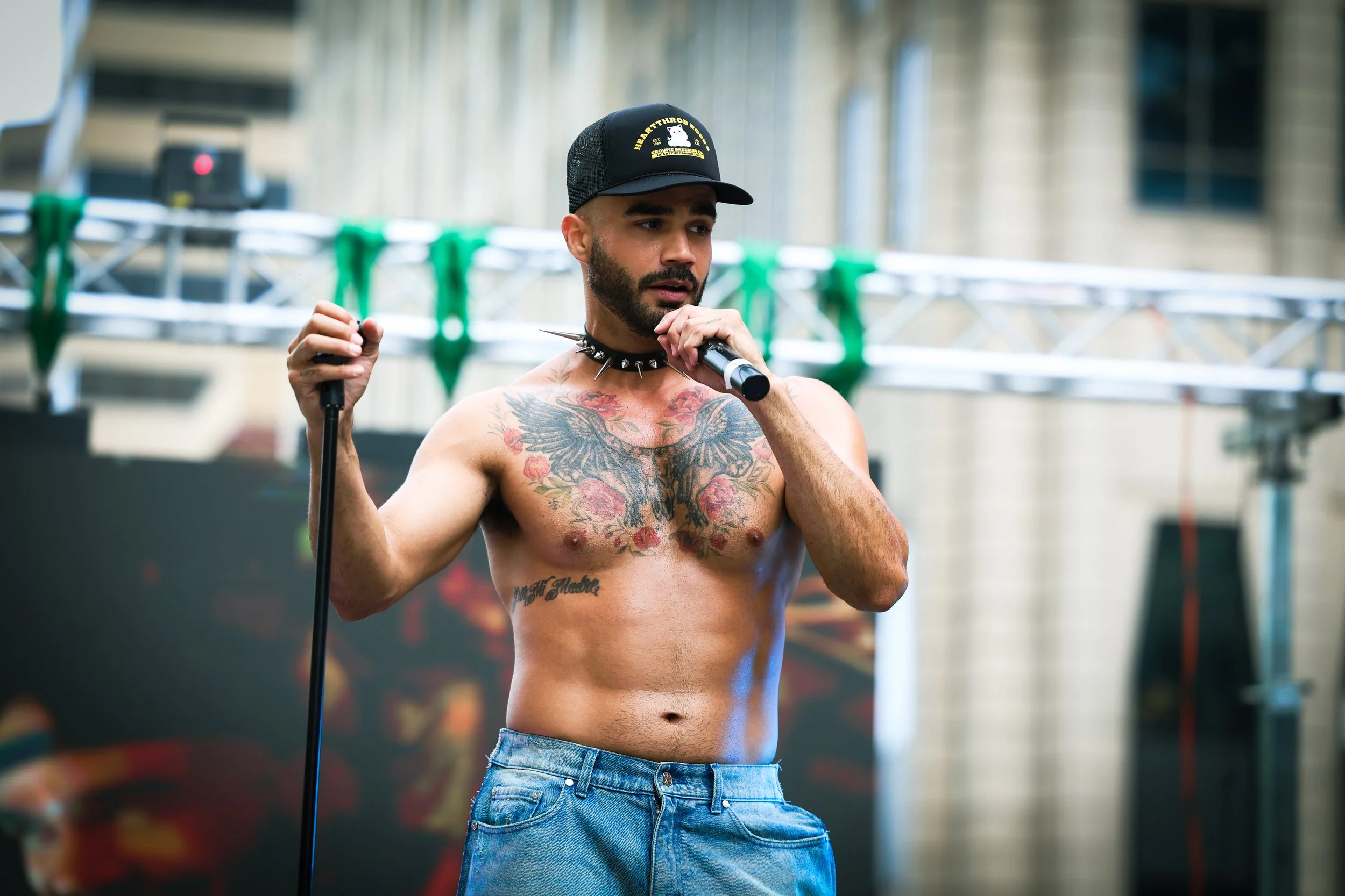 A shirtless man with tattoos, wearing a baseball cap and a spiked choker, holds a microphone in one hand and a stick in the other, standing on a stage with an urban backdrop.
