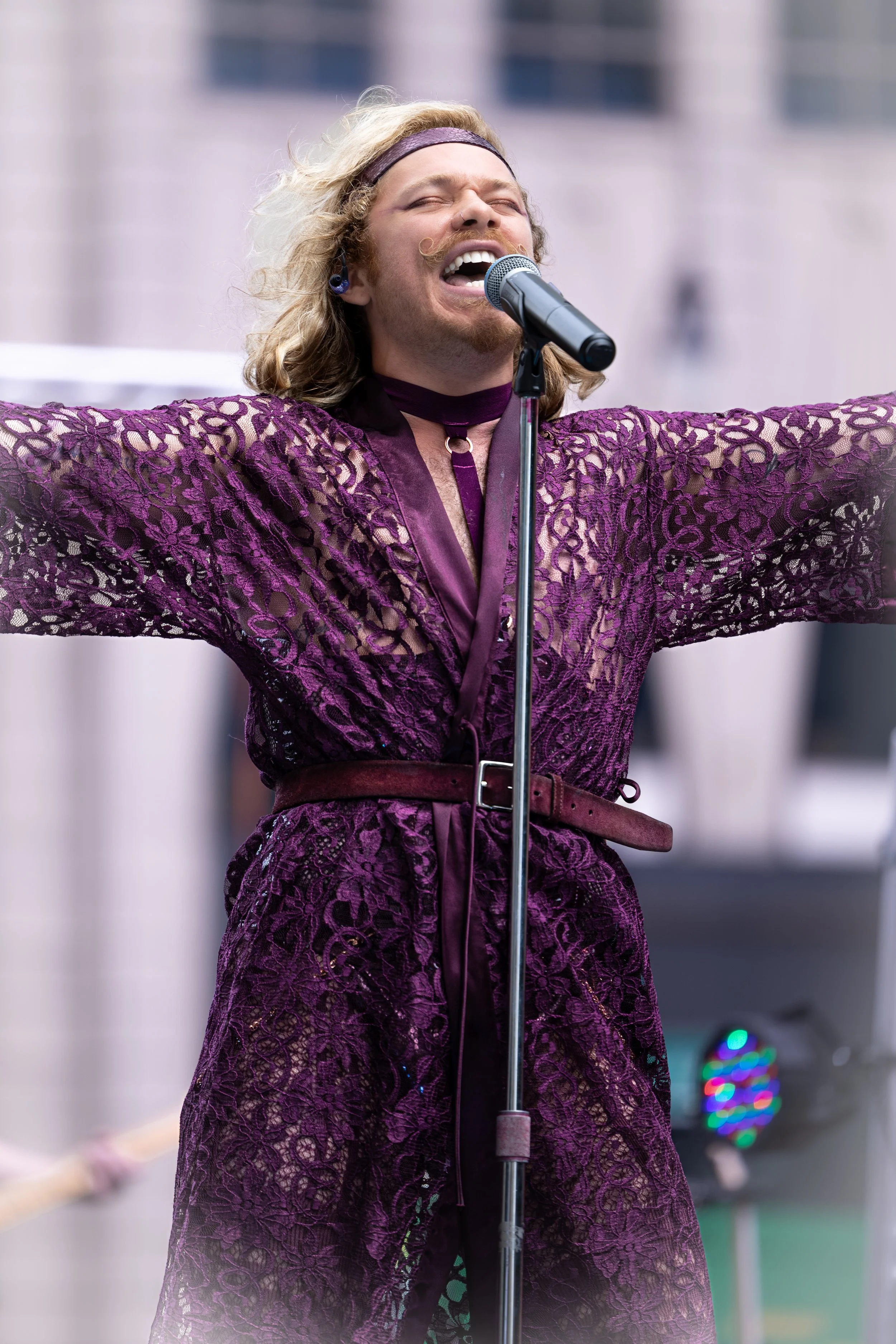 A man with long, curly blonde hair, wearing a lavender headband and a purple lace robe, passionately singing into a microphone with arms spread wide.