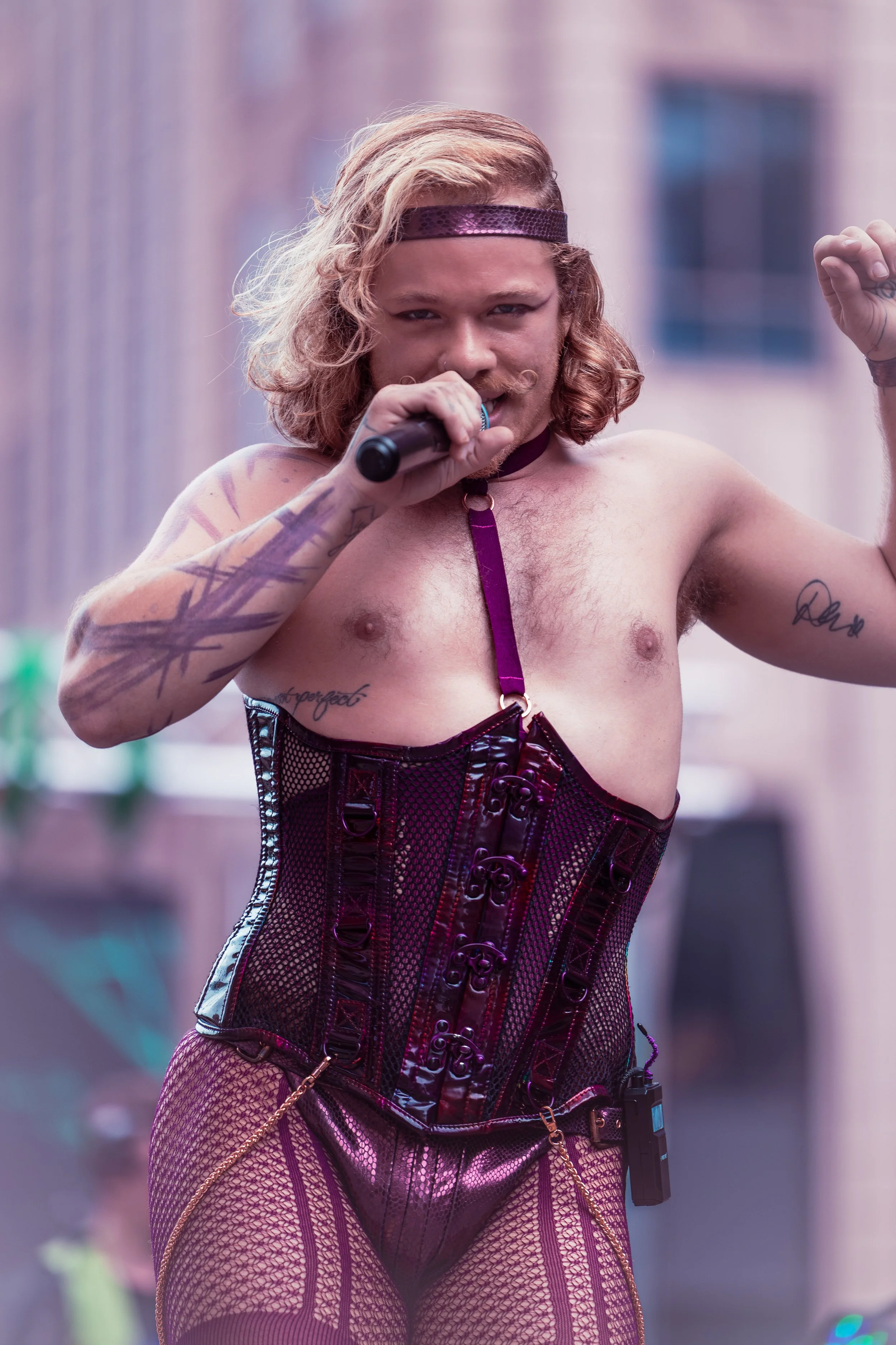 Person with long hair singing or speaking into a microphone, wearing a shiny purple corset and fishnet stockings, with tattoos on arms, outdoors in an urban area.