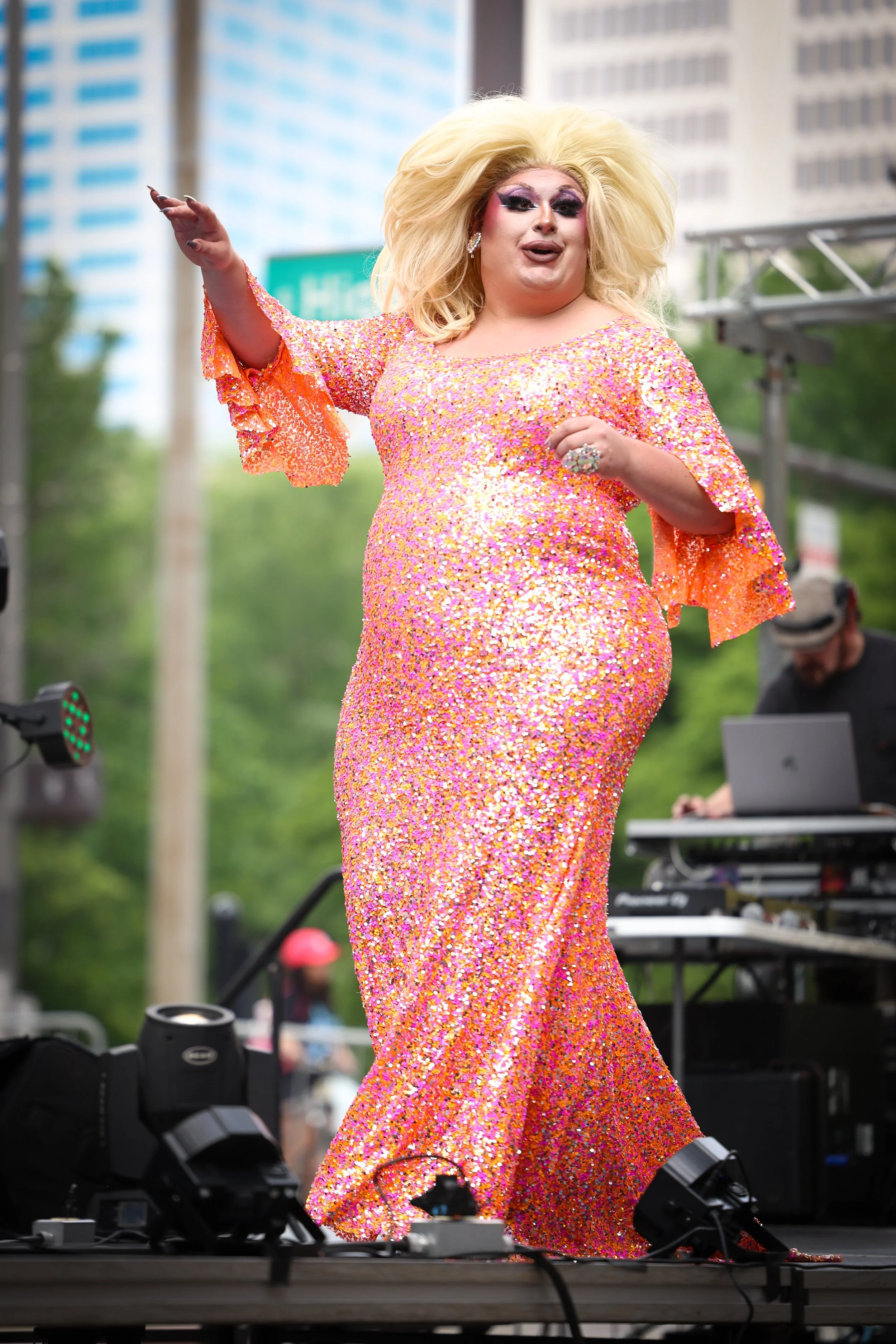 A drag queen performing on stage wearing a long, pink and orange sequined dress with bell sleeves, with voluminous blonde hair and dramatic makeup.