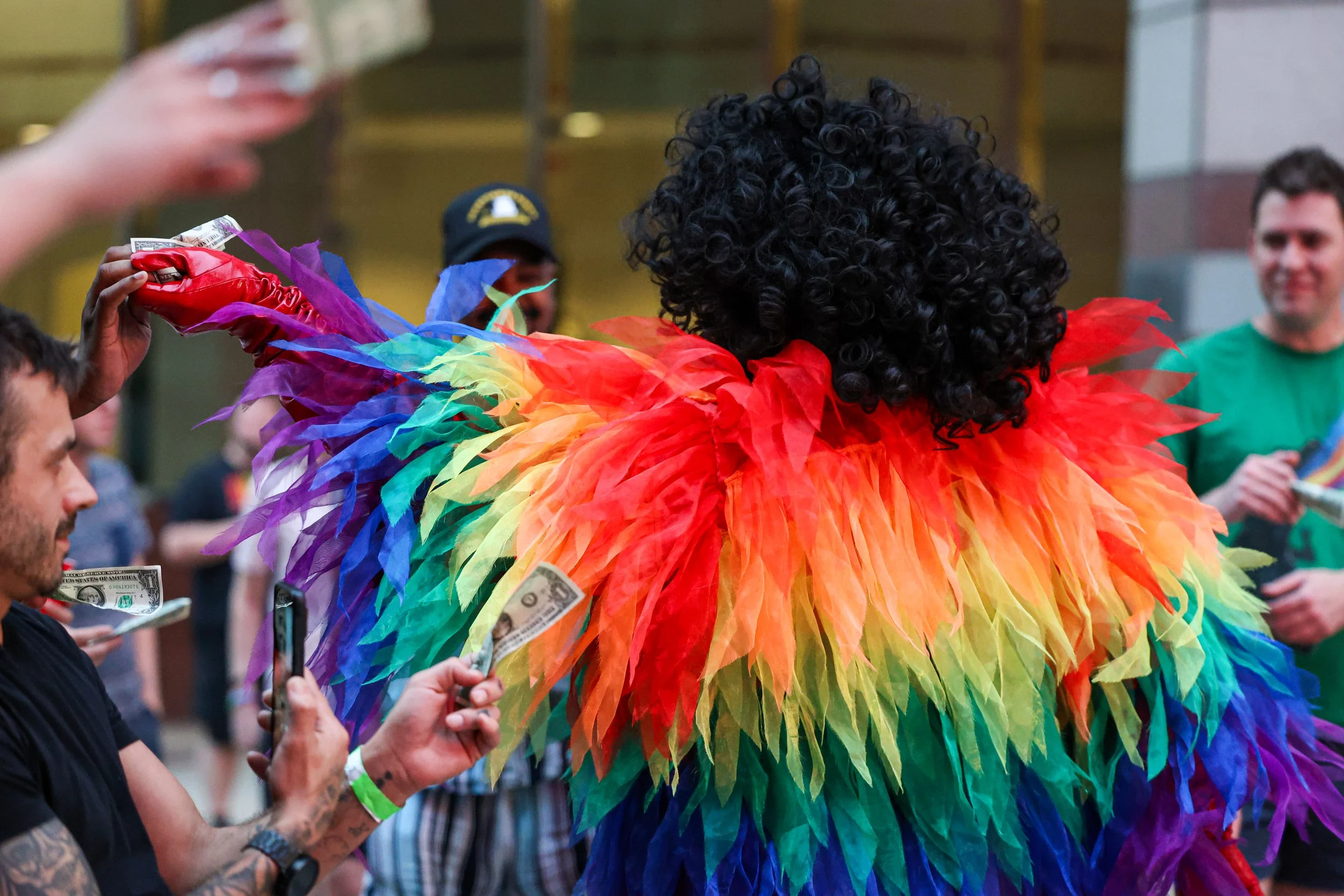 Person wearing a rainbow-colored feathered costume at a celebration, surrounded by people, some holding money and taking photos.